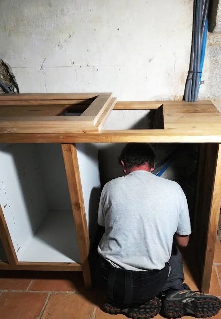 A man working on installing or repairing a wooden cabinet under a countertop in a room with unfinished walls.
