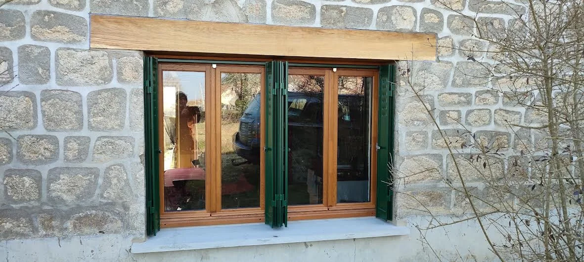A stone house exterior with a large wooden-framed glass window and green shutters, surrounded by a leafless tree branch.