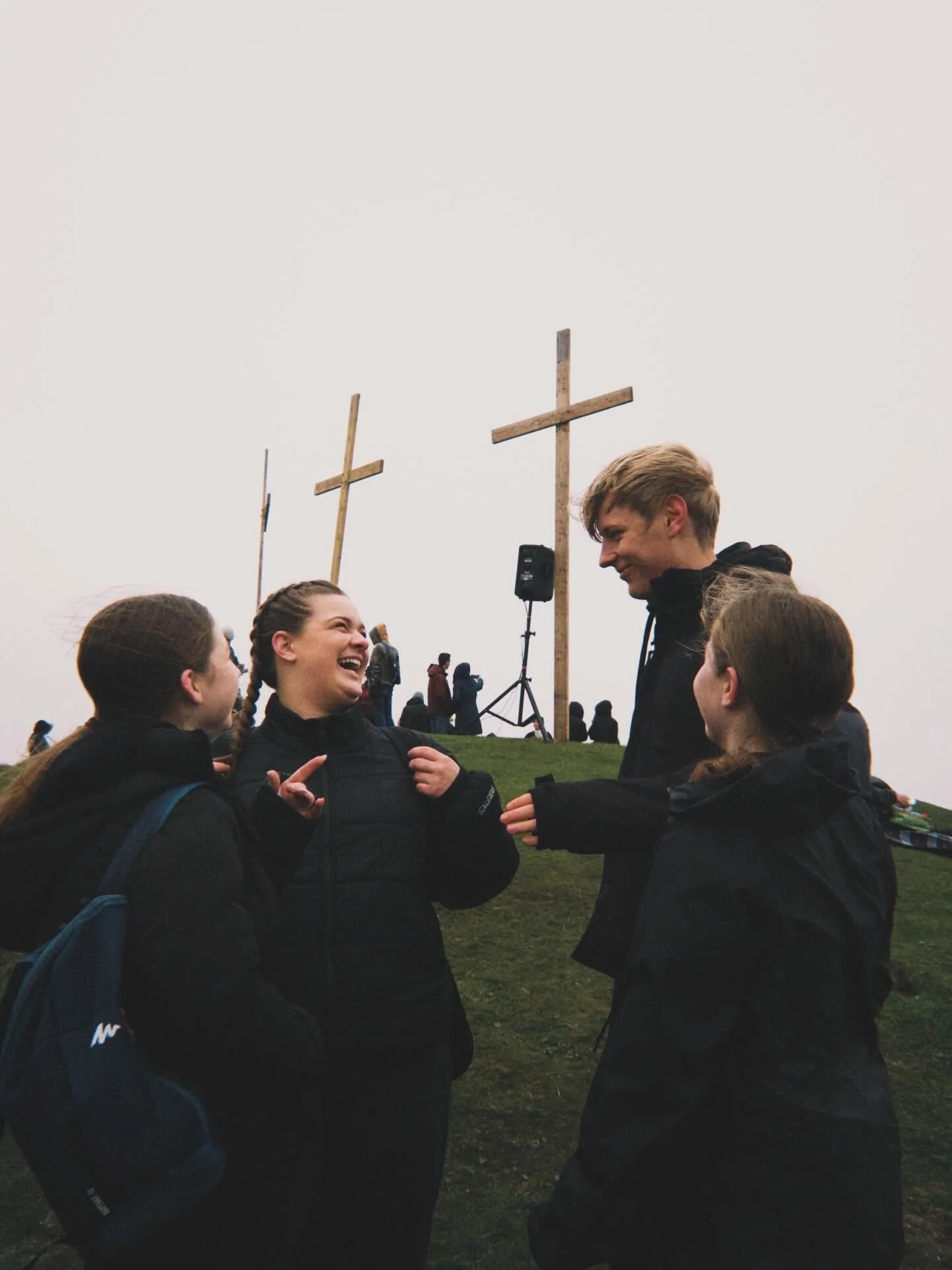 Group of five young people, four women and one man, smiling and talking outdoors on an overcast day, with three wooden crosses in the background.