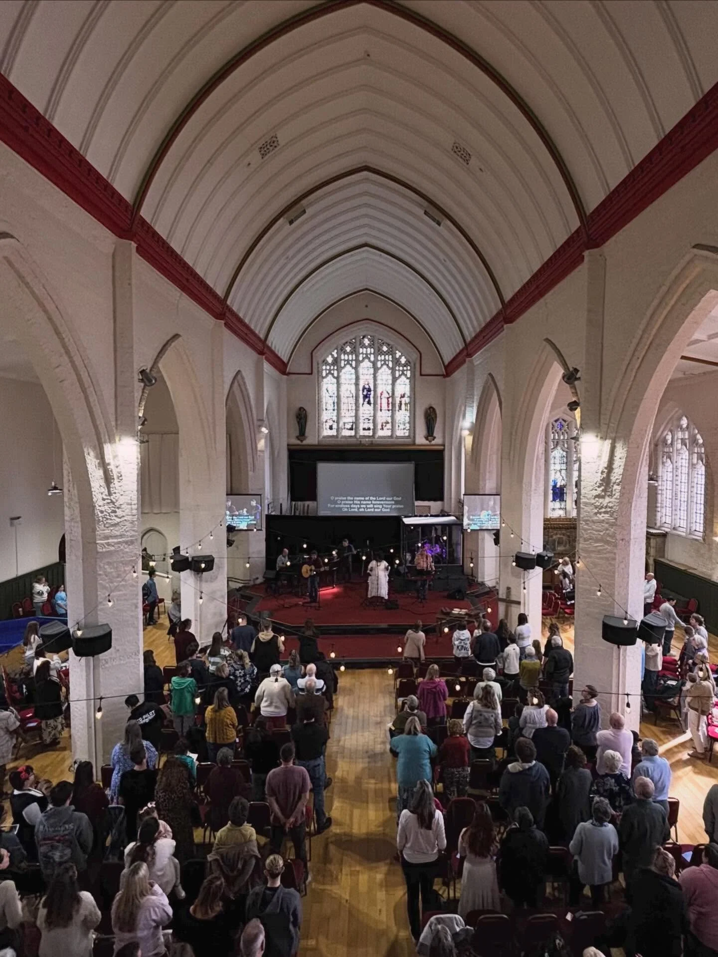 People gather in a church with high arched ceilings, stained glass windows, and a stage where a band is performing, for a religious service.