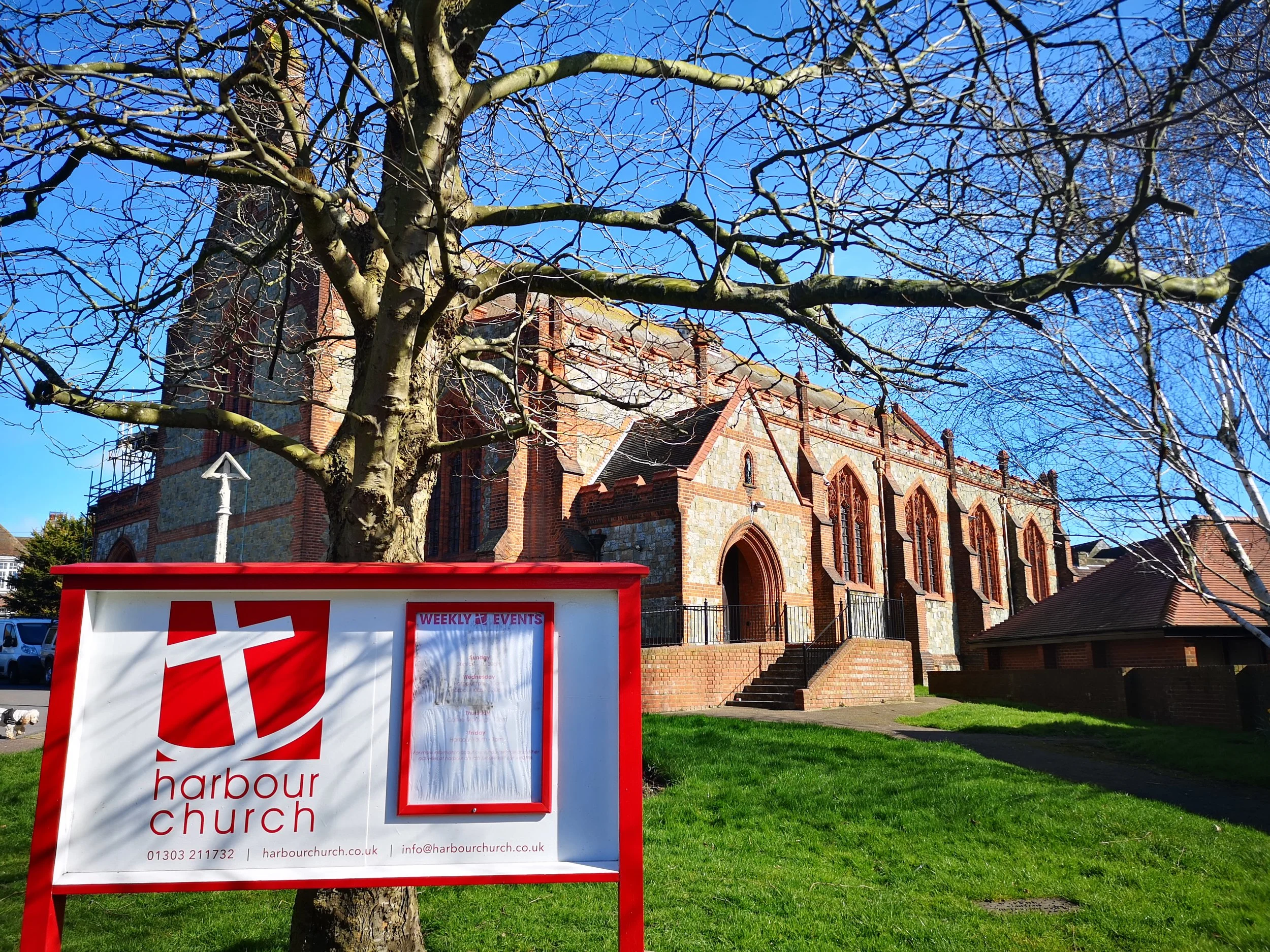 A red and white sign for Harbour Church in front of a leafless tree and a red brick church building with arched windows and a steep roof under a clear blue sky.