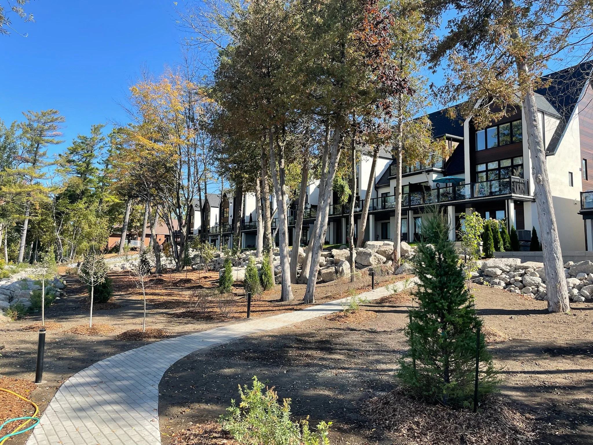 Residential building with large windows and balconies, surrounded by trees with autumn foliage, gravel and paved pathways in a landscaped yard.