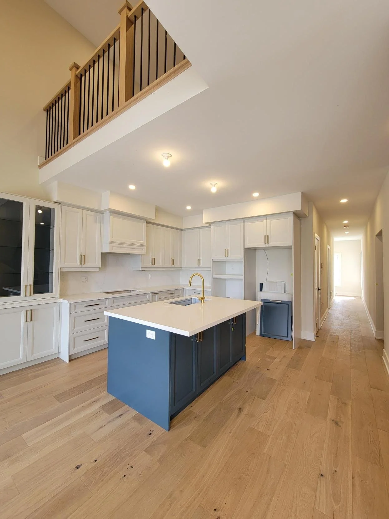 Interior of a modern kitchen with white cabinets, a navy blue island with a gold faucet, and hardwood flooring. There is an upper level balcony with wooden and black metal railing visible from below.