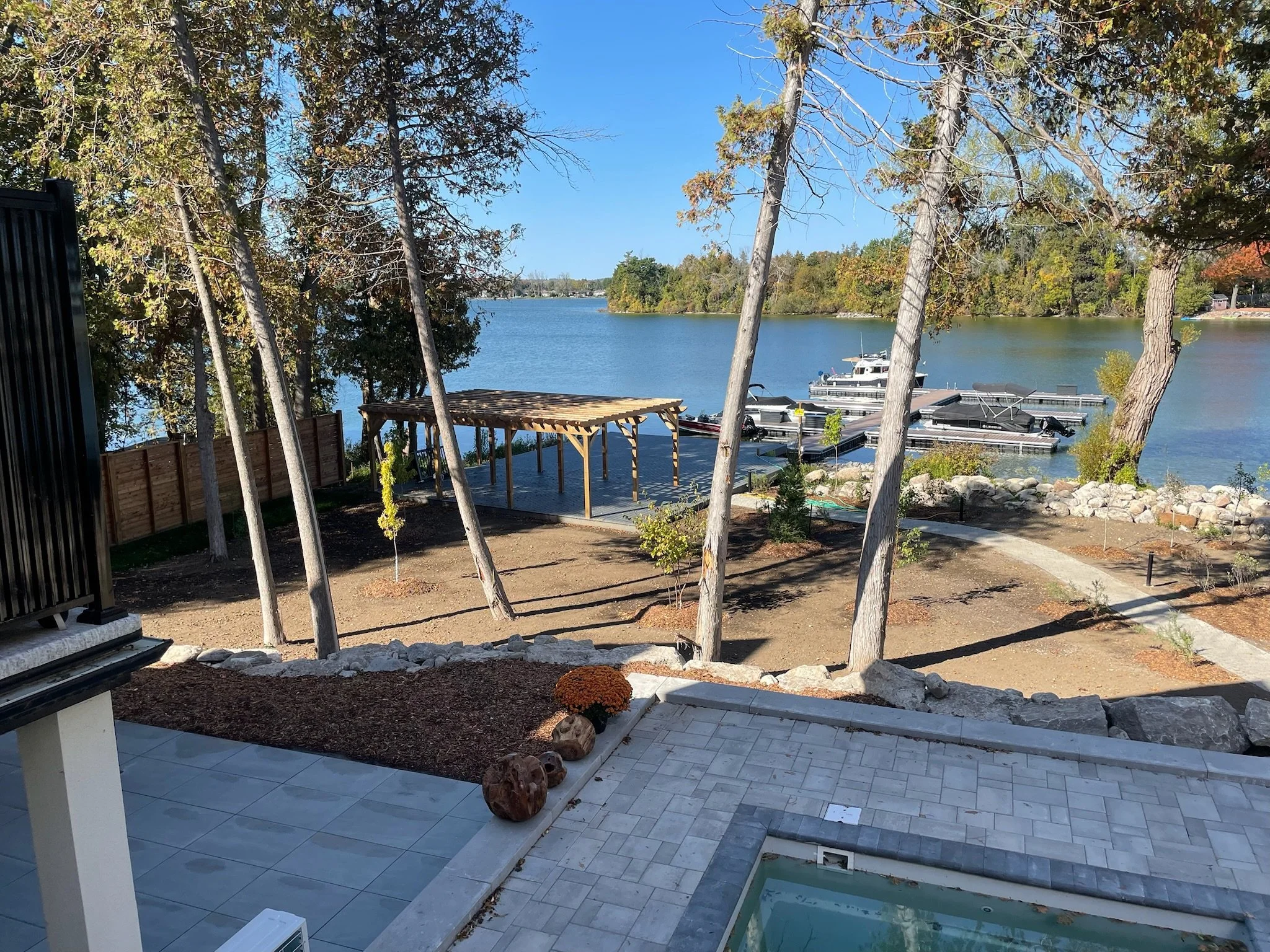 View of a lake with boats docked at a marina, trees with autumn foliage, and a partially constructed or renovated outdoor area with a tiled patio, wooden deck, and decorative rocks.