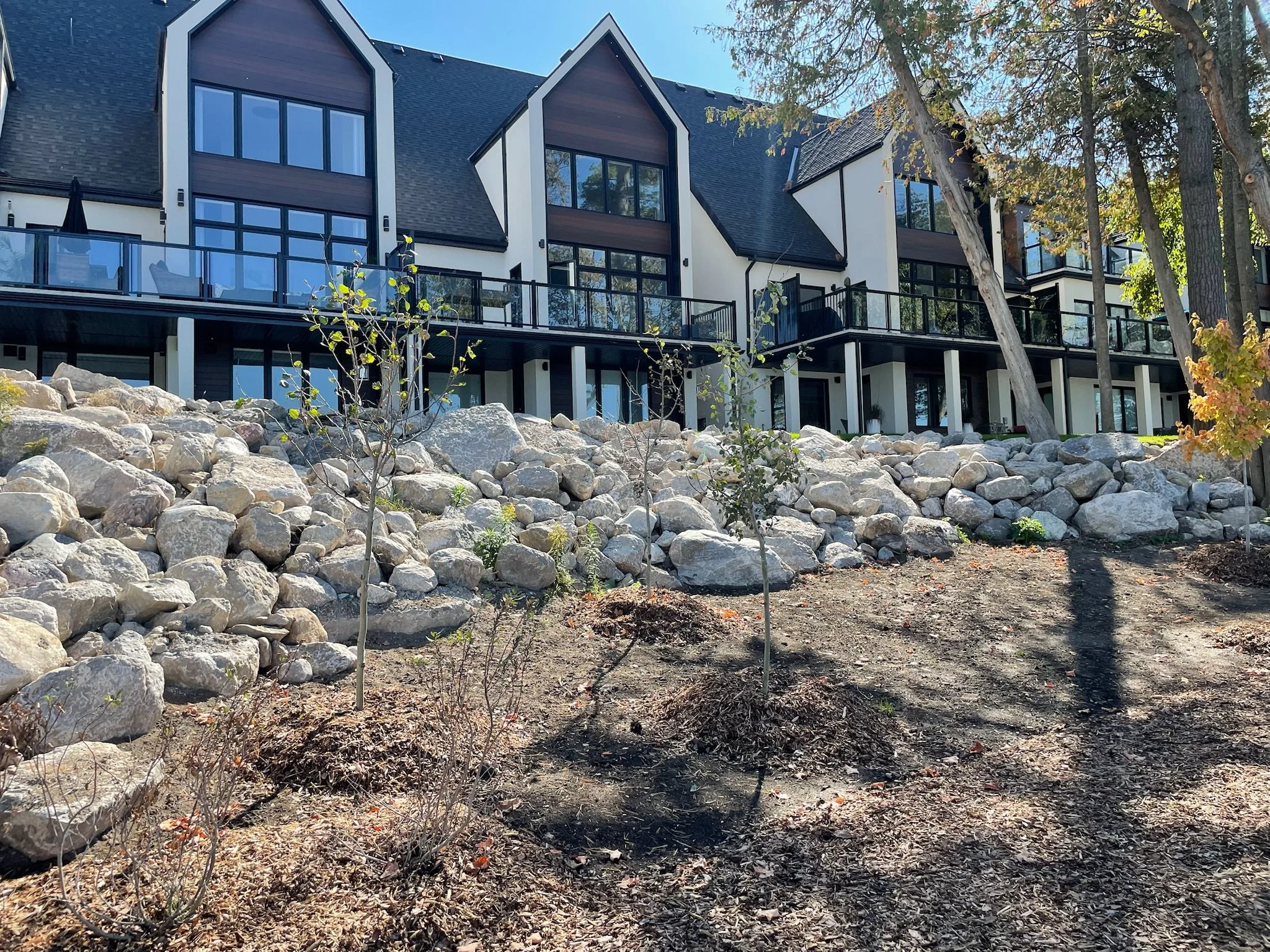 View of a modern multi-story residential building with large windows and balconies, behind a rocky landscape with small plants and trees, under a clear blue sky.