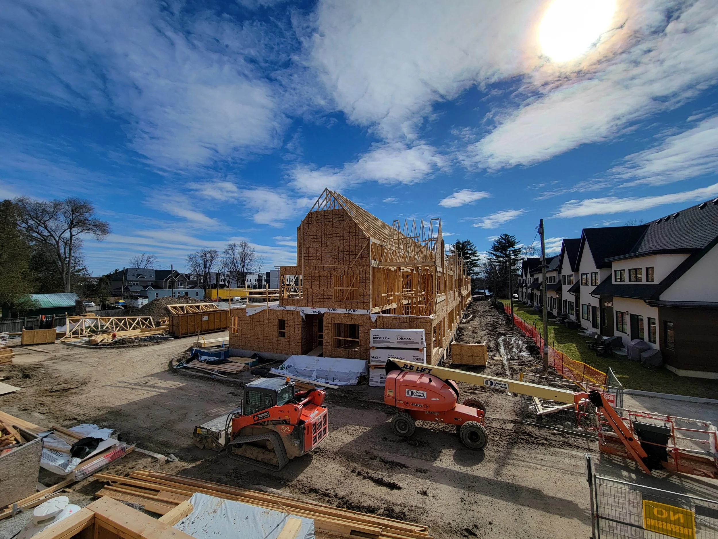 Construction site with wooden framework for a new house, construction equipment, and nearby completed houses under a partly cloudy sky.