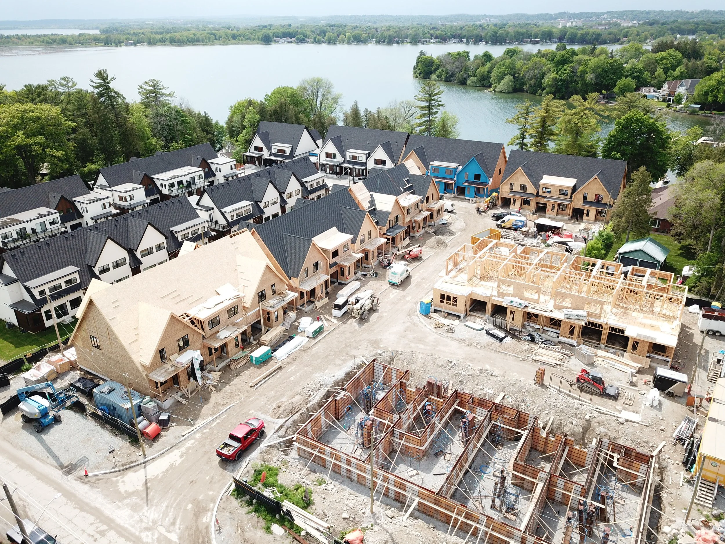 Aerial view of a construction site next to a lake with green trees and residential houses in the background.