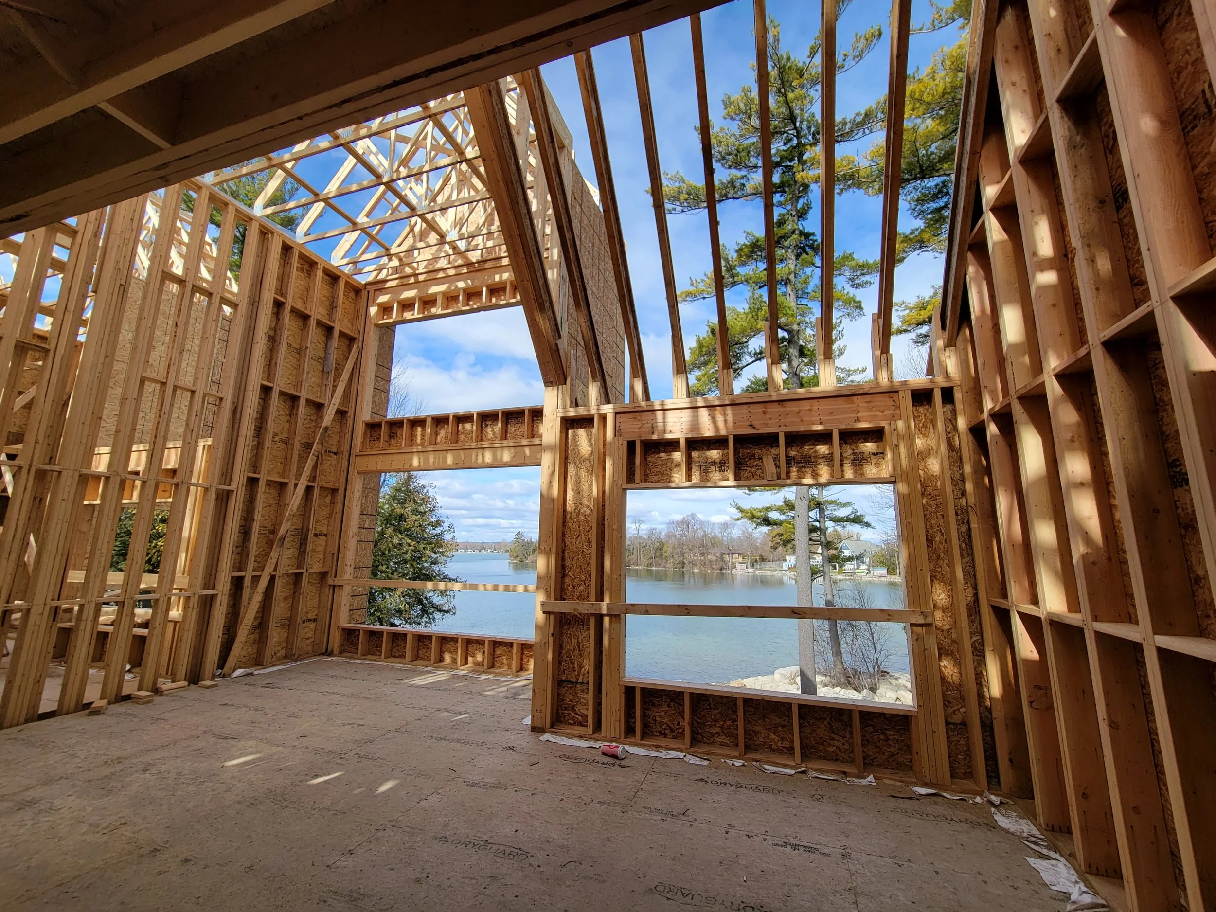 The view from an unfinished wooden house framing overlooking a lake and trees on a partly cloudy day.