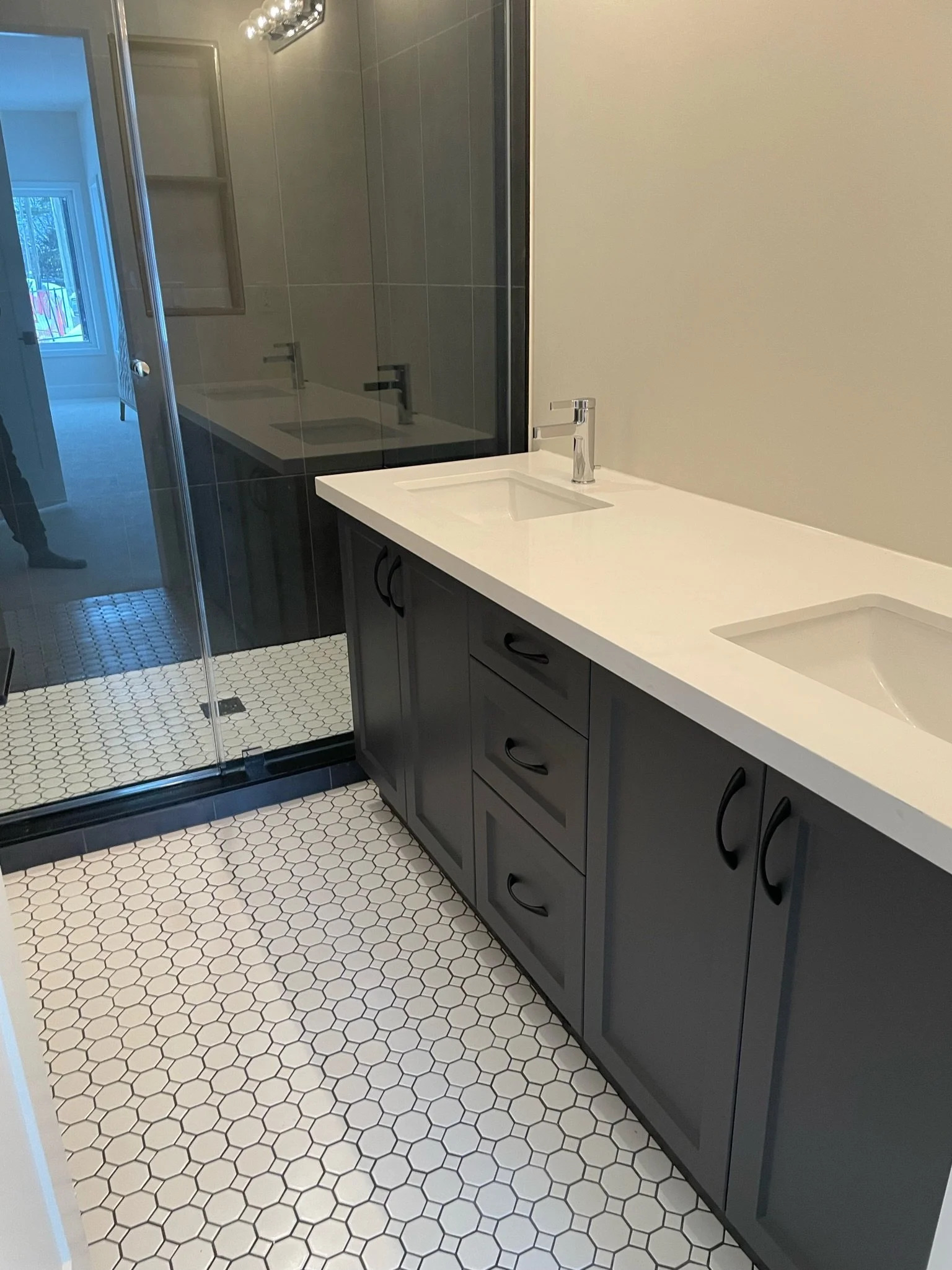 Bathroom with a white countertop and double sinks, dark gray cabinets, hexagonal white floor tiles, a walk-in shower with a glass door, a window, and a wall with beige paint.