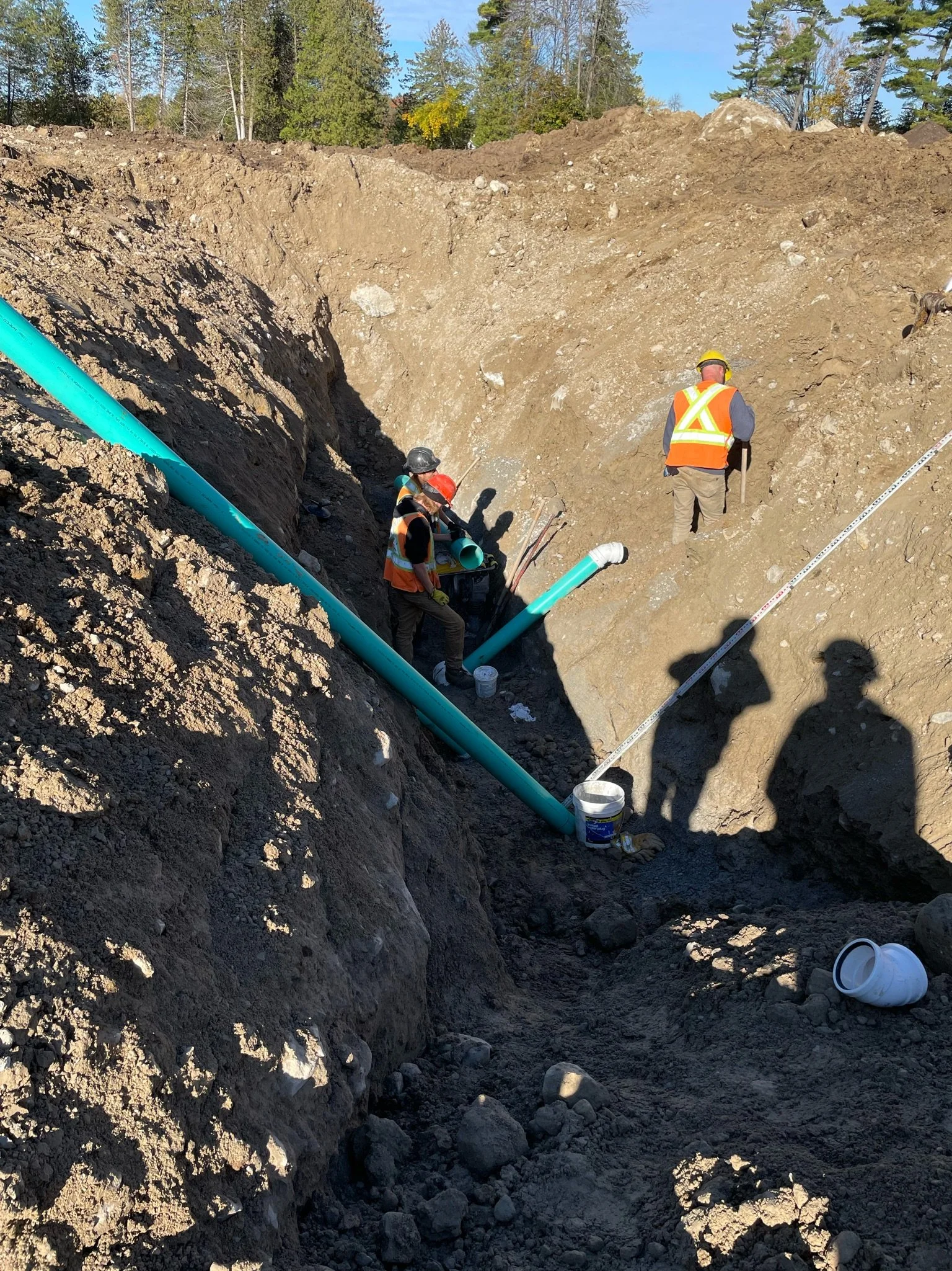 Construction workers in safety gear working underground on a trench with exposed blue pipes, dirt, and tools, with trees and blue sky in the background.