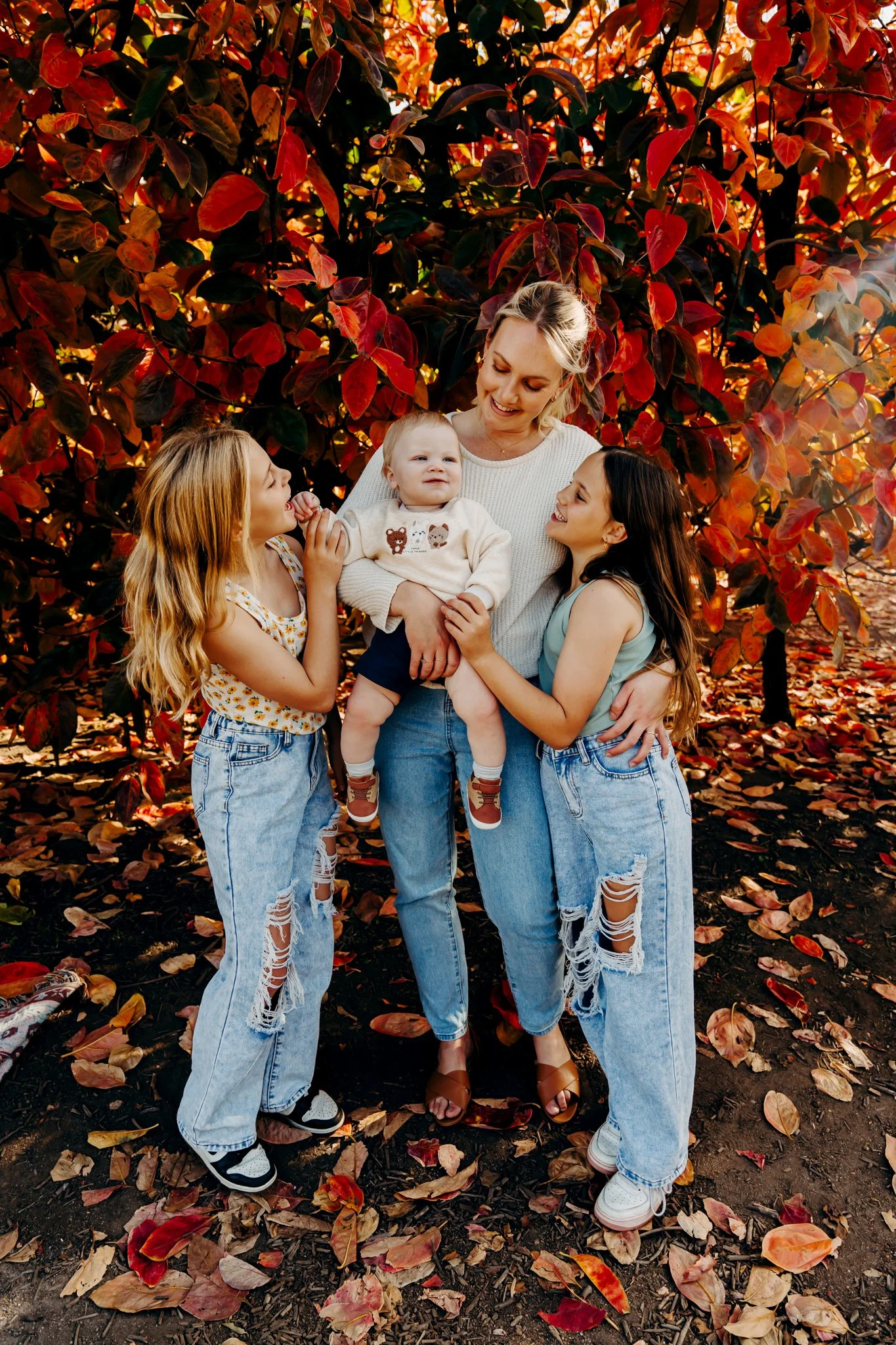 A woman and three children standing outdoors in front of a large tree with red and orange autumn leaves, with fallen leaves on the ground.