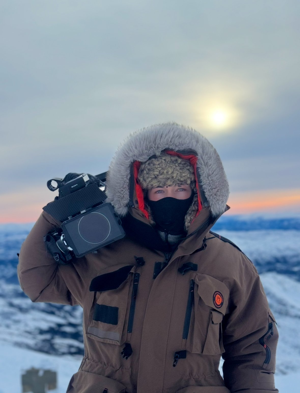 Person wearing a heavy winter coat with a large fur-lined hood, face mask, and furry hat, carrying an electronic device, outdoors in a snowy landscape during sunset.