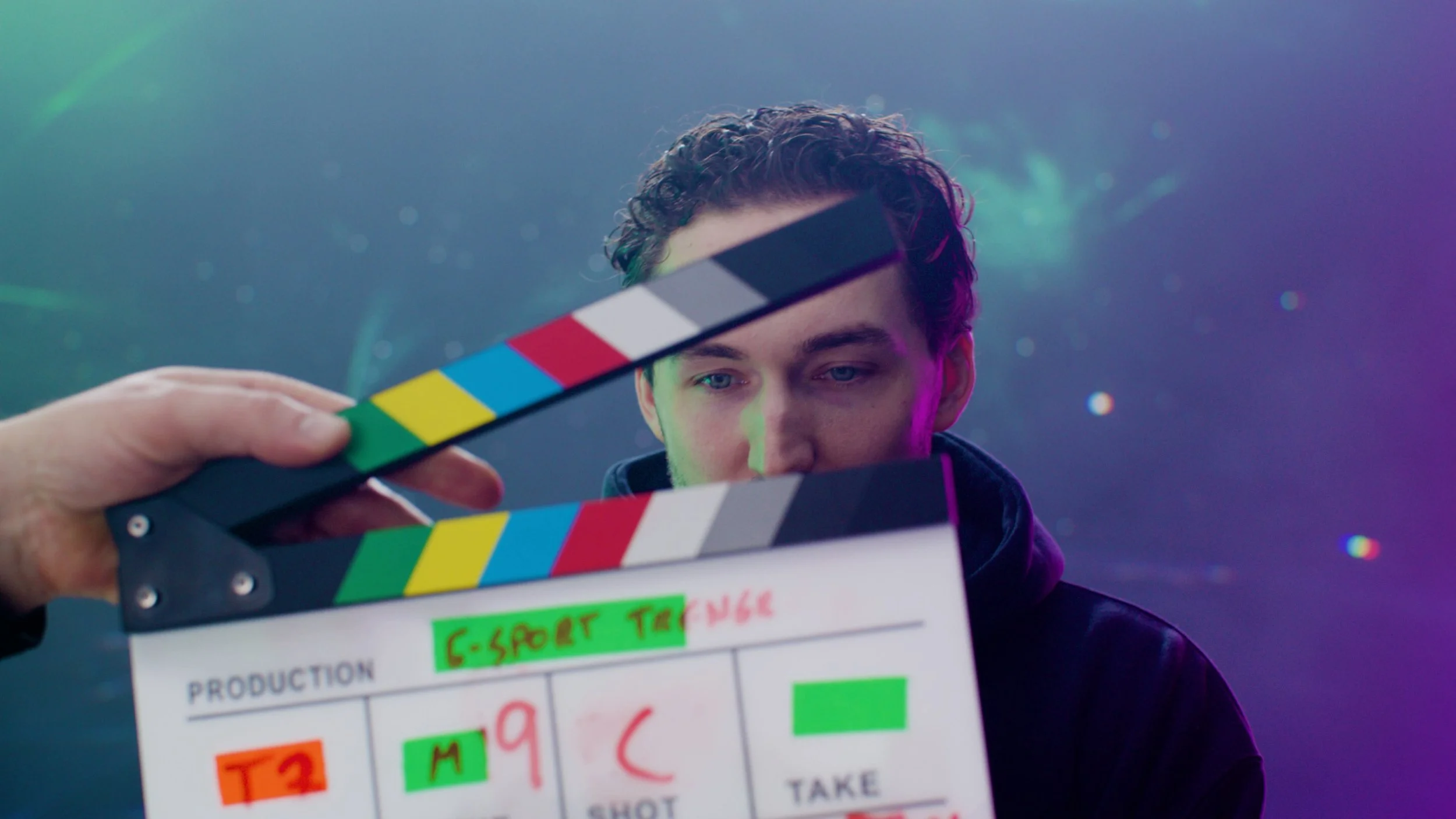 A person holding a clapperboard on a film set, with a young man in the background looking at the camera and a colorful, dreamy backdrop.
