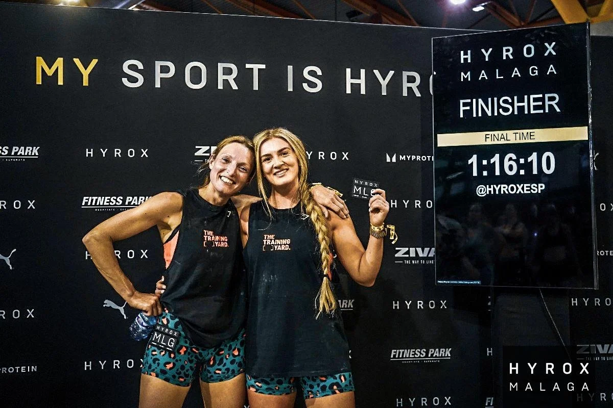 Two women standing together, smiling, one holding a finisher card after a race, with a backdrop and digital sign displaying the race completion time of 1:16:10 at Hyrox Malaga.