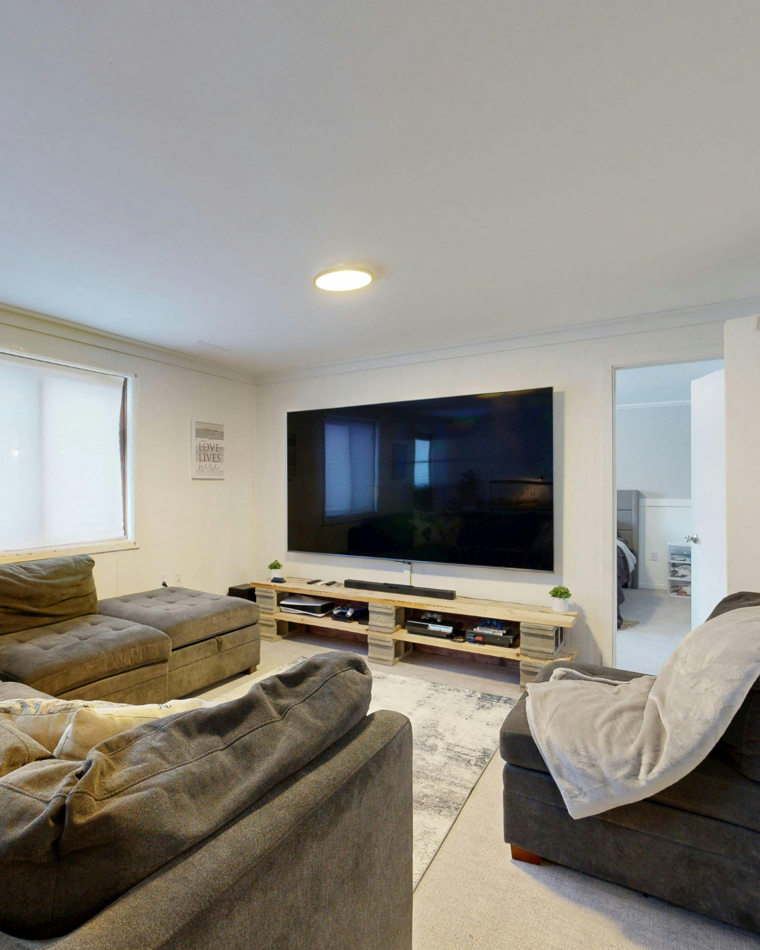 Living room with beige couches, a large flat-screen TV on a wooden stand, a window with white blinds, and a bedroom visible through an open door. Installation completed by Sound Select AV.