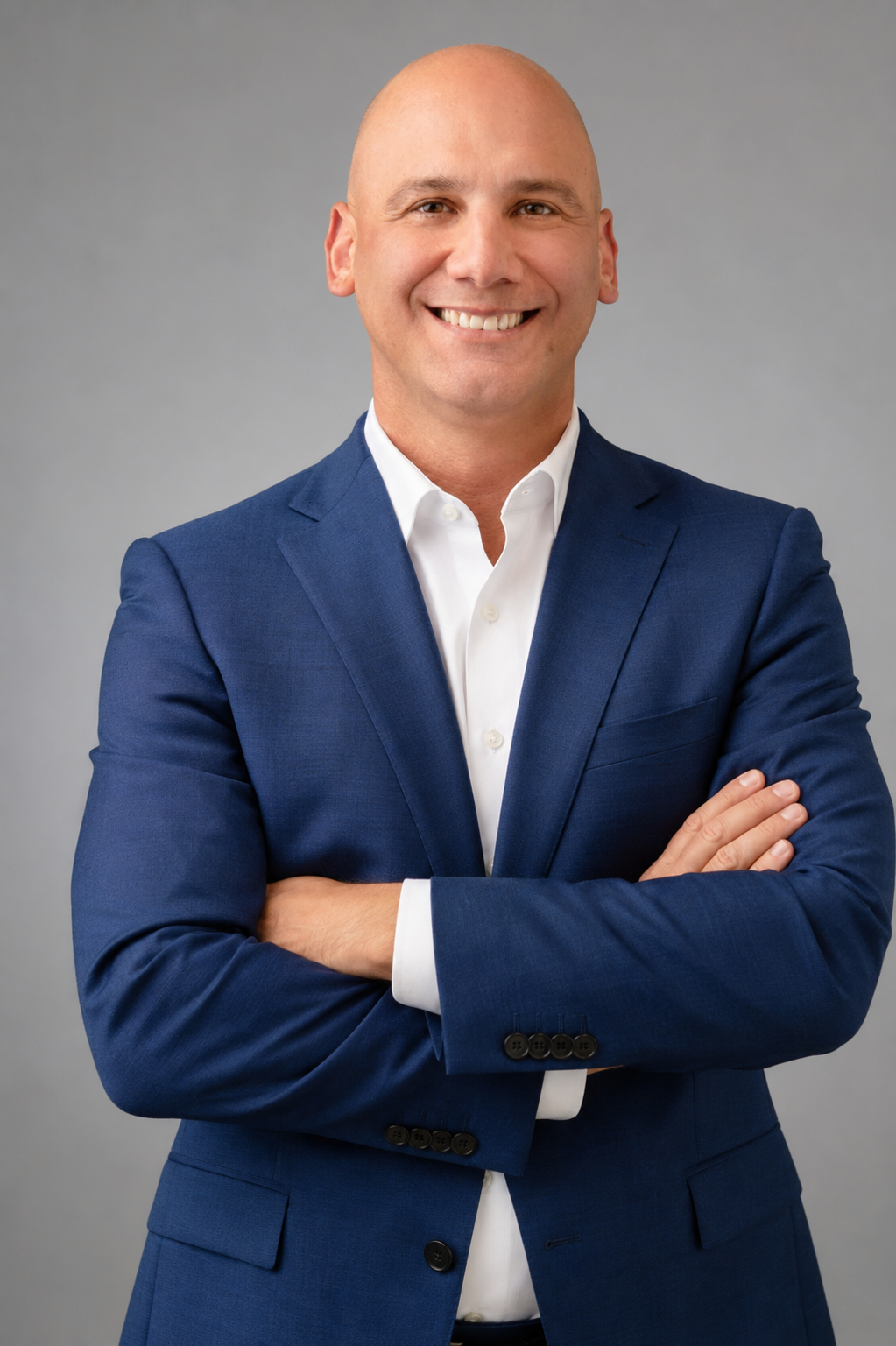 A smiling man with a bald head, wearing a blue suit and white shirt, standing with arms crossed against a neutral gray background.