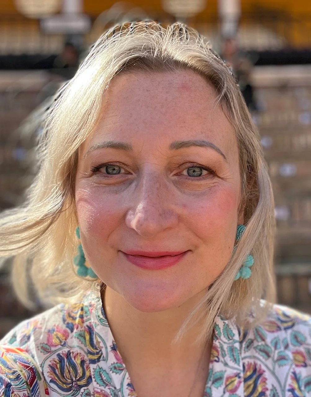 Close-up of a woman with blonde hair, blue eyes, and turquoise earrings, smiling in sunlight.