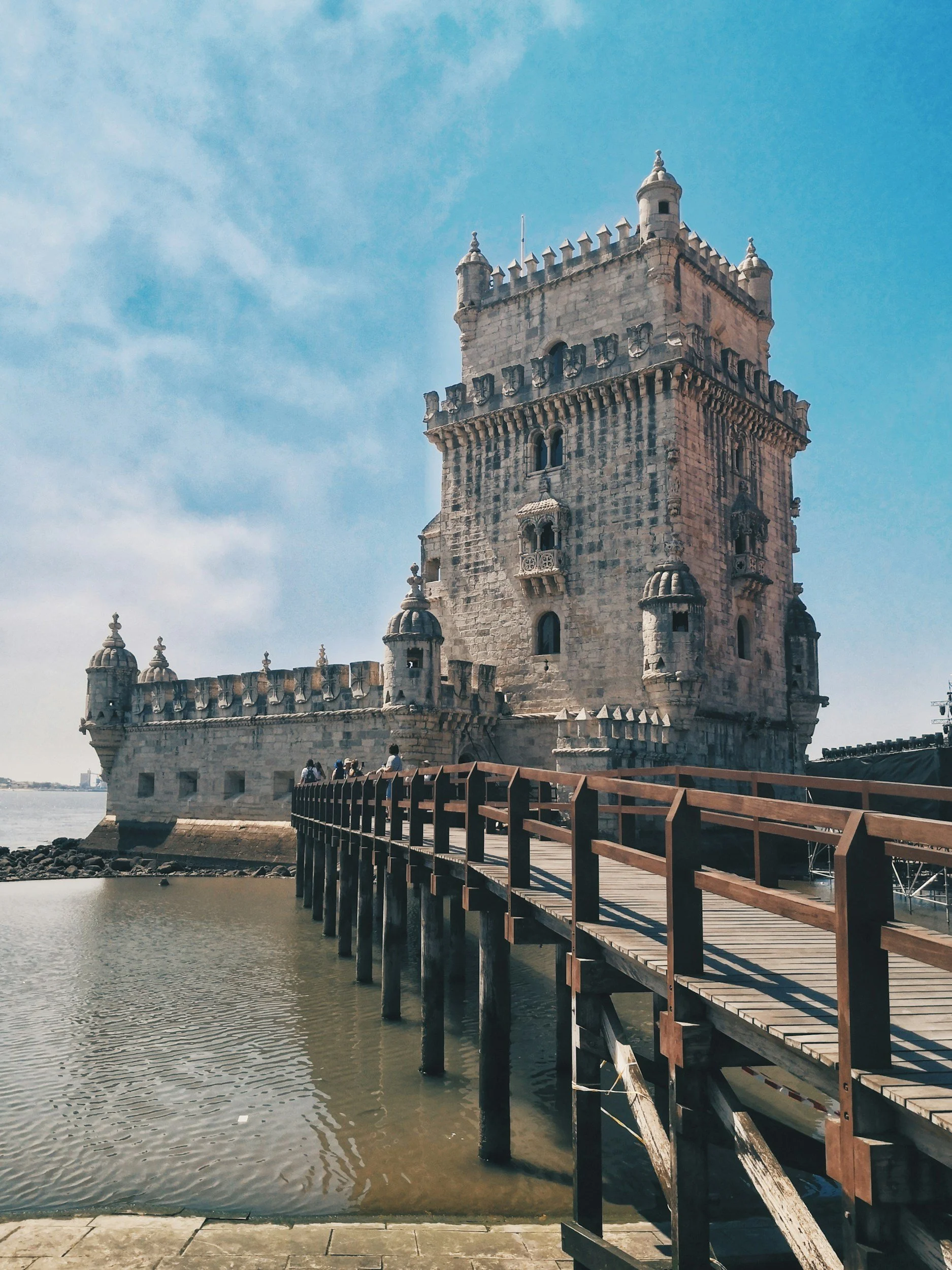 The historic Belém Tower, a fortress with turrets and battlements, located by the river with a wooden walkway leading to it, under a partly cloudy sky.