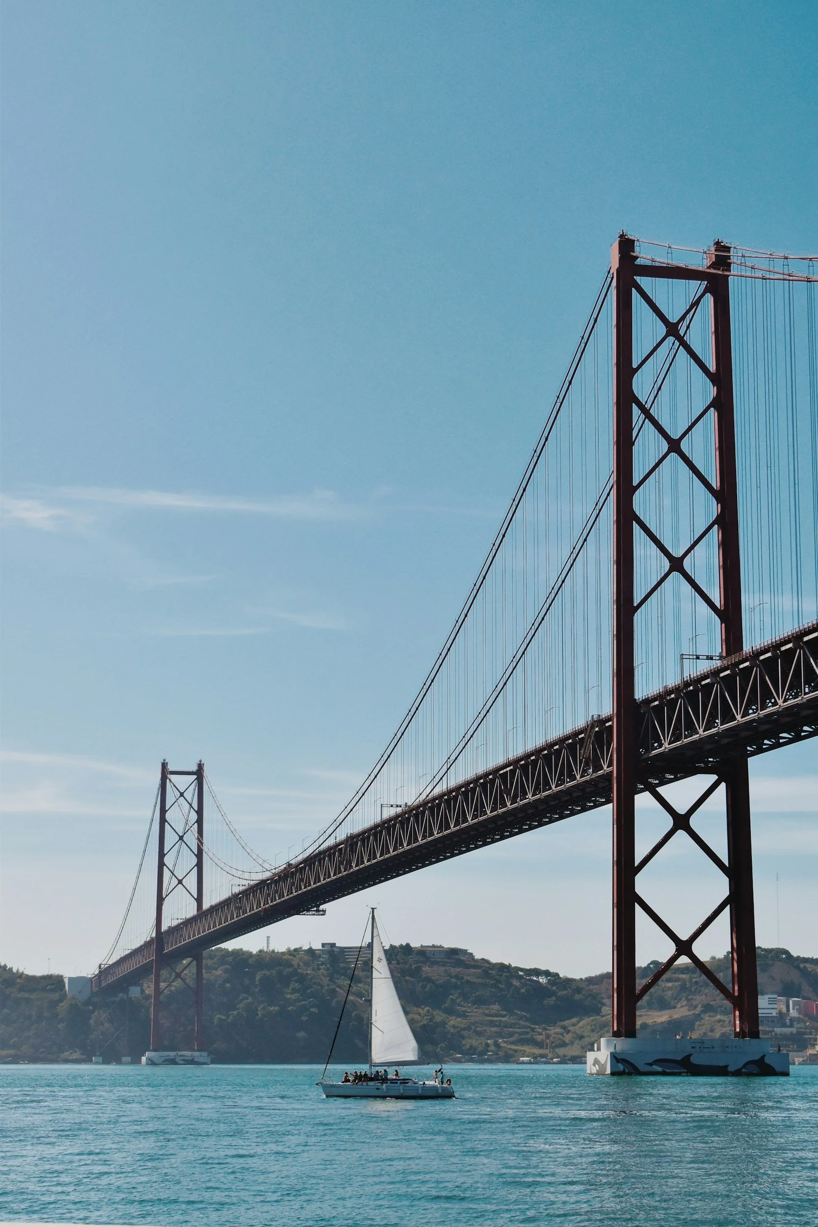 A sailboat on the water under a large red suspension bridge with a blue sky in the background.