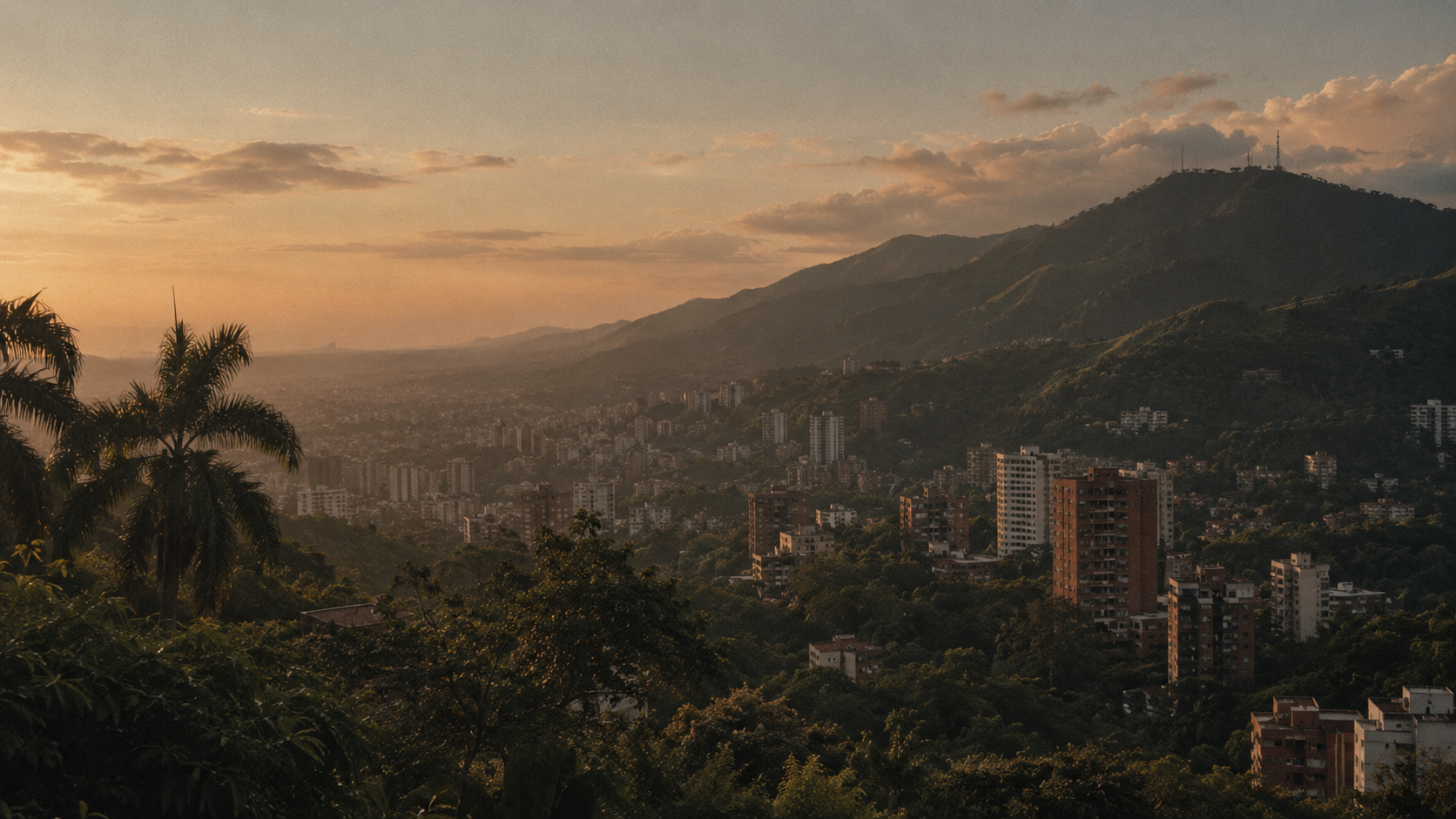Cityscape of a hilly urban area during sunset with tall buildings and lush green trees, mountains in the background, and a partly cloudy sky.