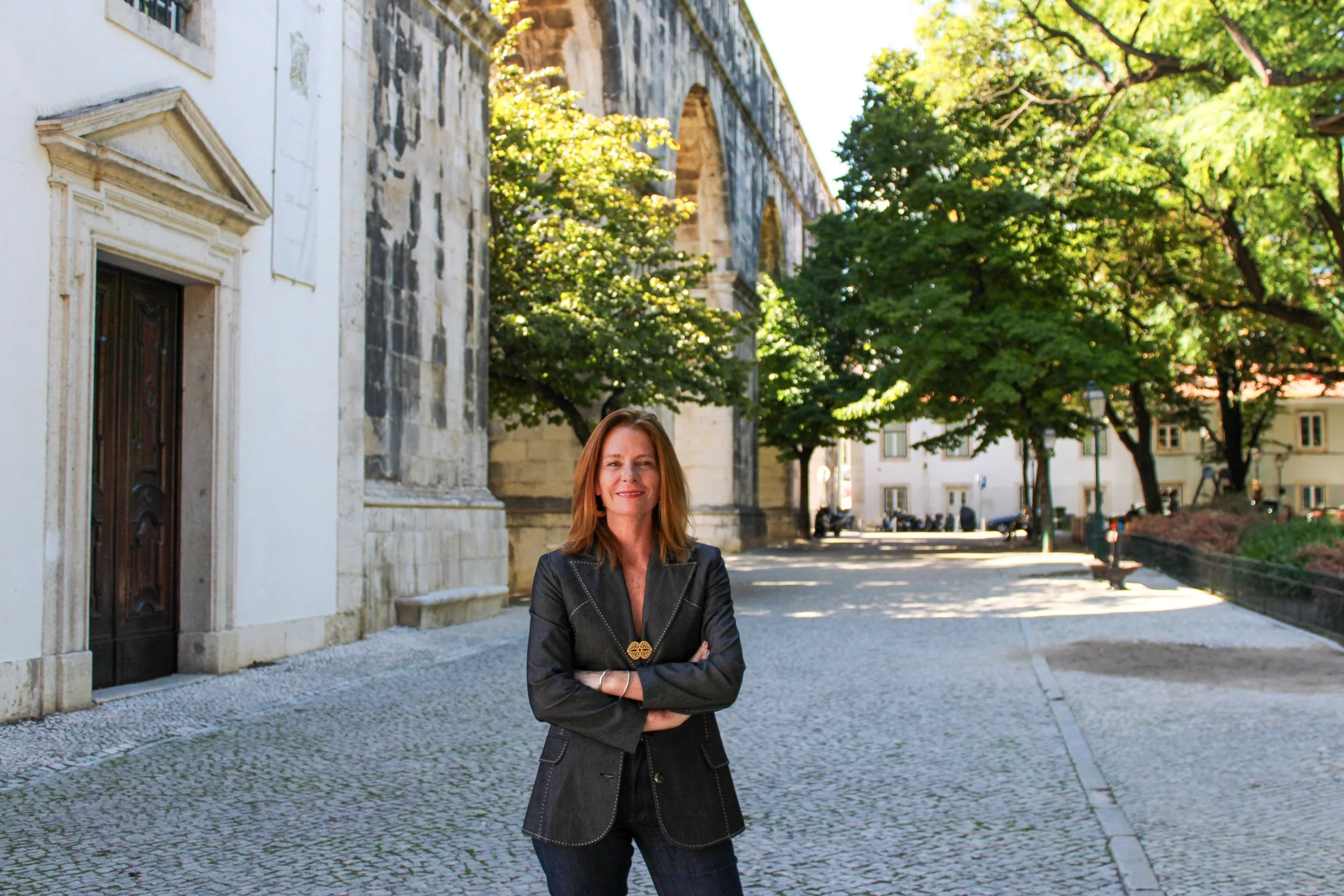 A woman with reddish-brown hair in a black jacket smiling with her arms crossed on a cobblestone street lined with trees and historic buildings.