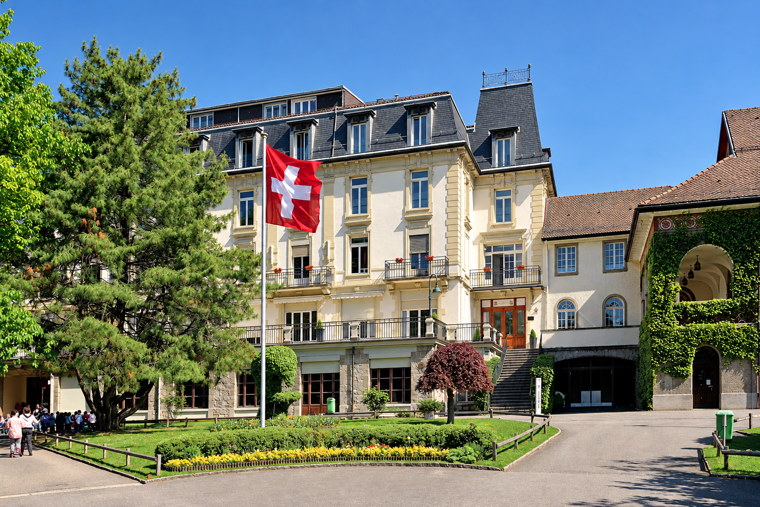 A large, elegant white building with multiple stories, surrounded by green trees and a well-maintained garden, with a Swiss flag on a flagpole in front.