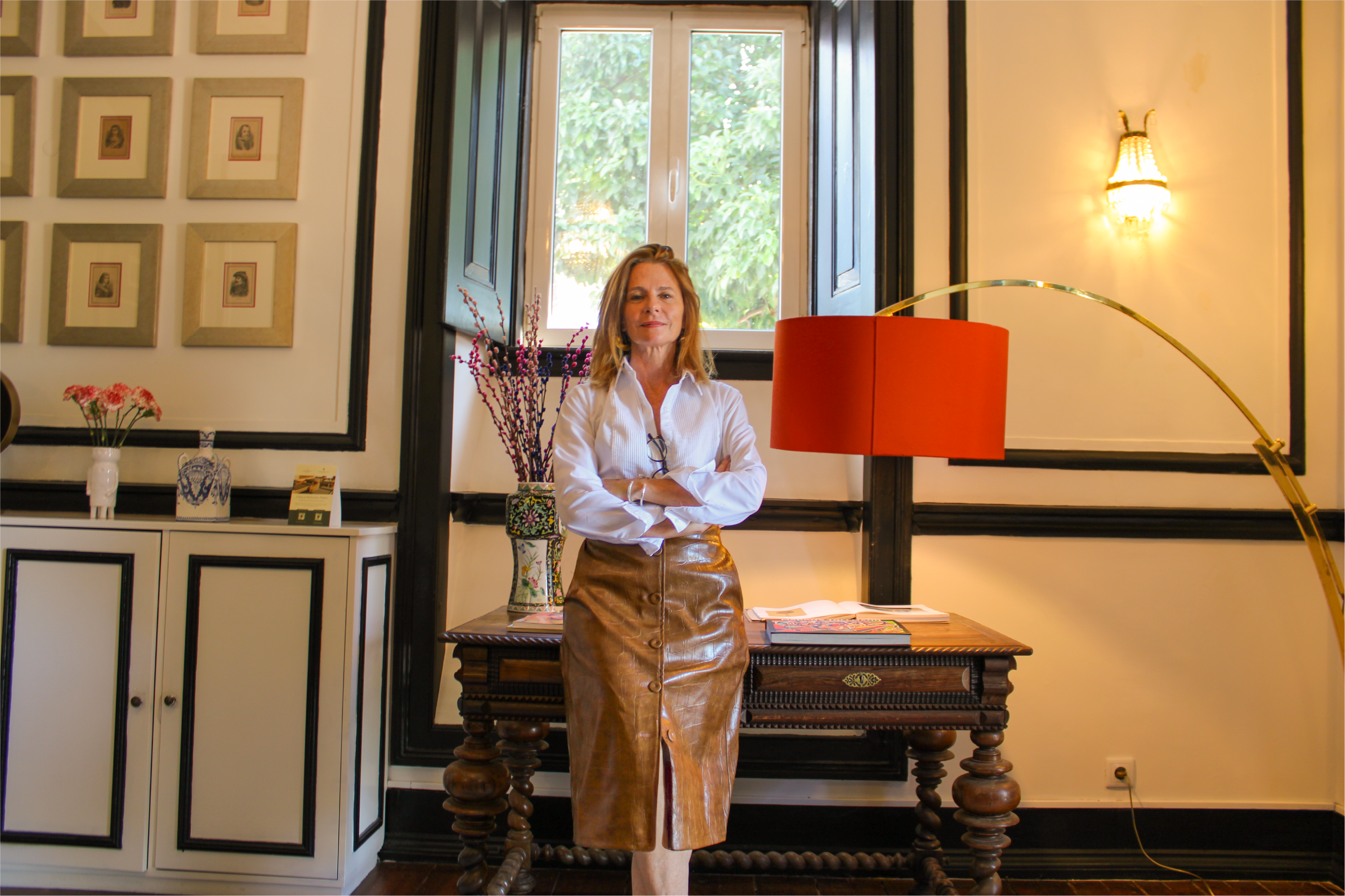 A woman with shoulder-length hair, wearing a white blouse and a leather skirt, standing with her arms crossed in a warmly lit room decorated with framed pictures, potted plants, and a piano.