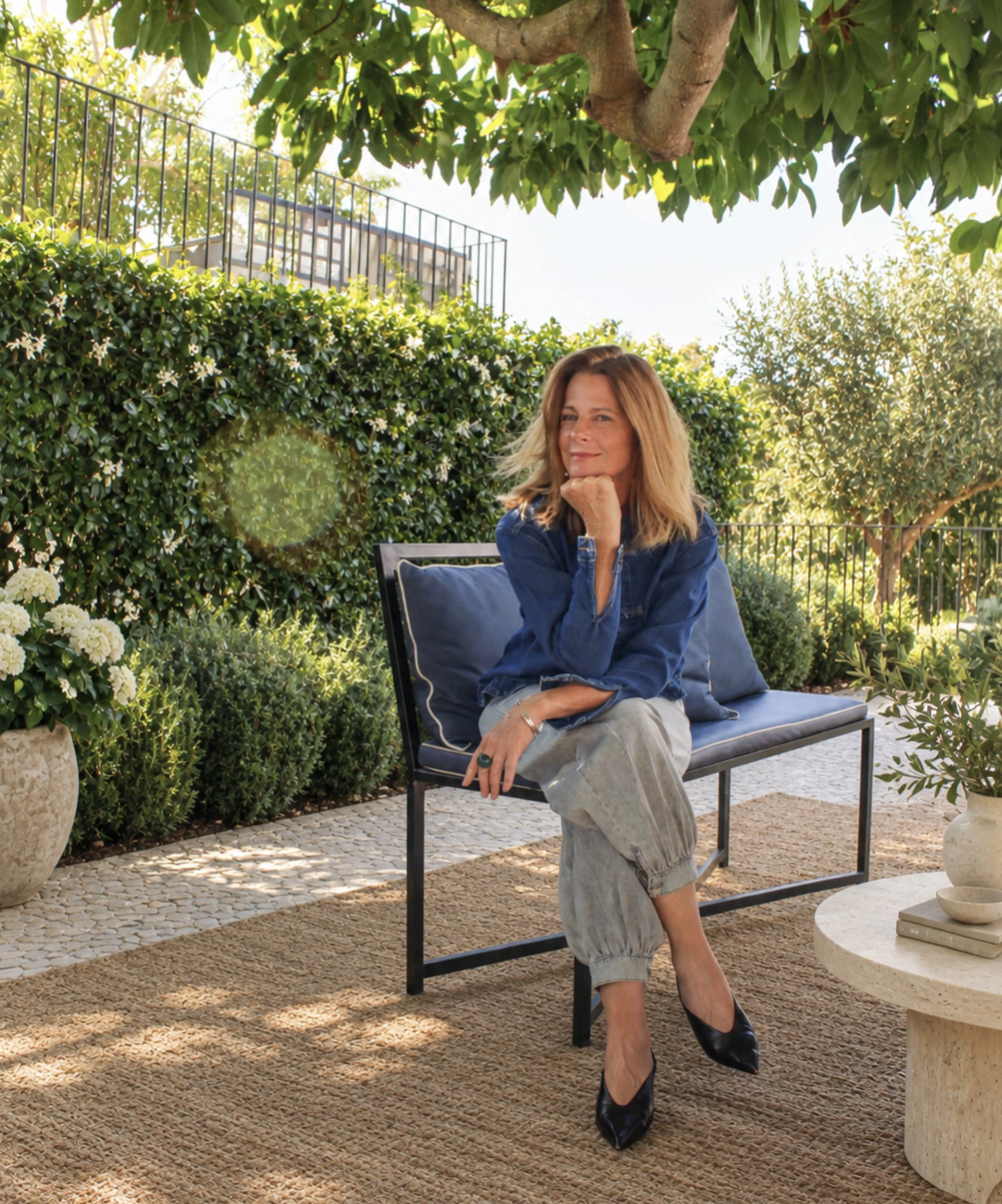 Woman sitting on a black outdoor chair with her chin resting on her hand in a garden setting with lush green foliage, potted plants, and a beige stone table, wearing a blue denim shirt, gray pants, and black heels, under a tree with broad leaves and sunny weather.