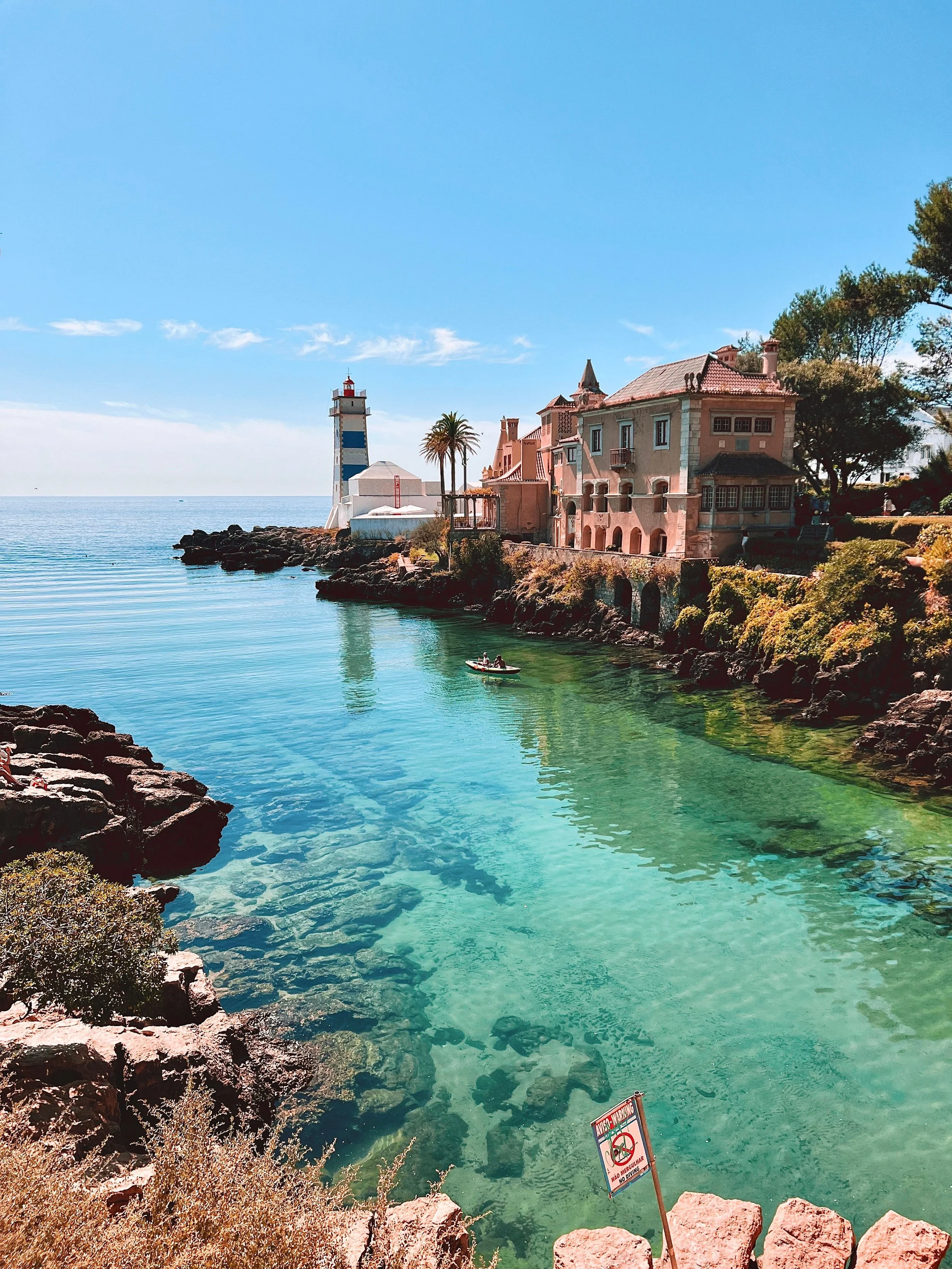 A coastal scene with clear turquoise water, rocky shoreline in the foreground, a pink building with a red-tiled roof along the water, a lighthouse with blue and white stripes, and a boat with two people on the water. There are trees and a warning sign in the scene.