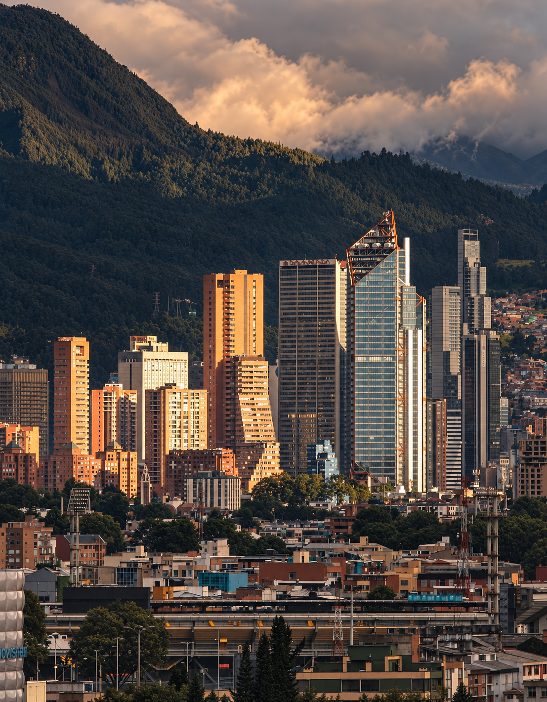A city skyline with tall modern skyscrapers set against green mountain slopes and partly cloudy sky at sunset.