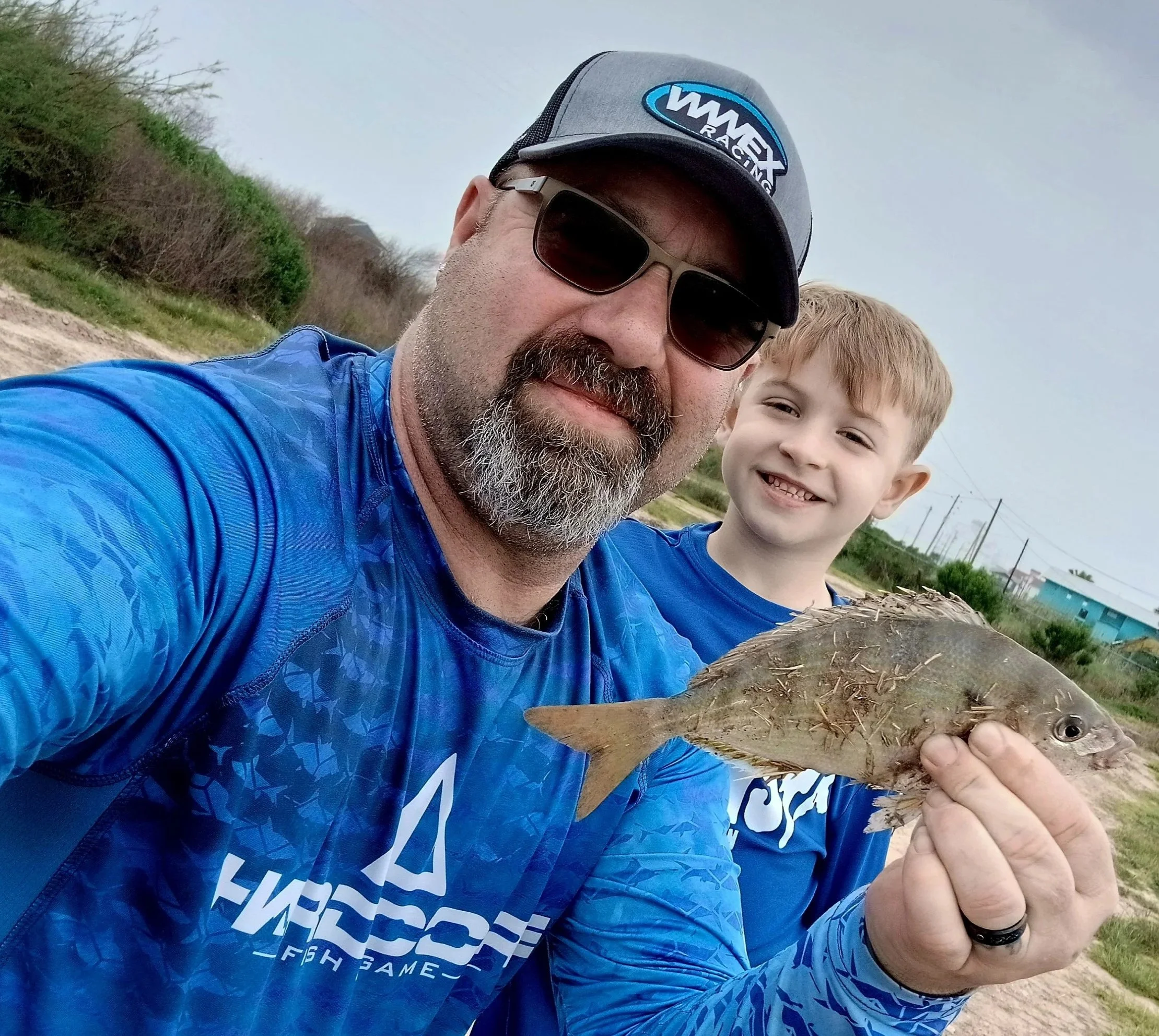 A man wearing sunglasses, a gray cap with a racing logo, and a blue shirt holds a fish, smiling at the camera. A young boy with light hair is also smiling in the background. They are outdoors in a rural area with dirt paths, green trees, and an overcast sky.