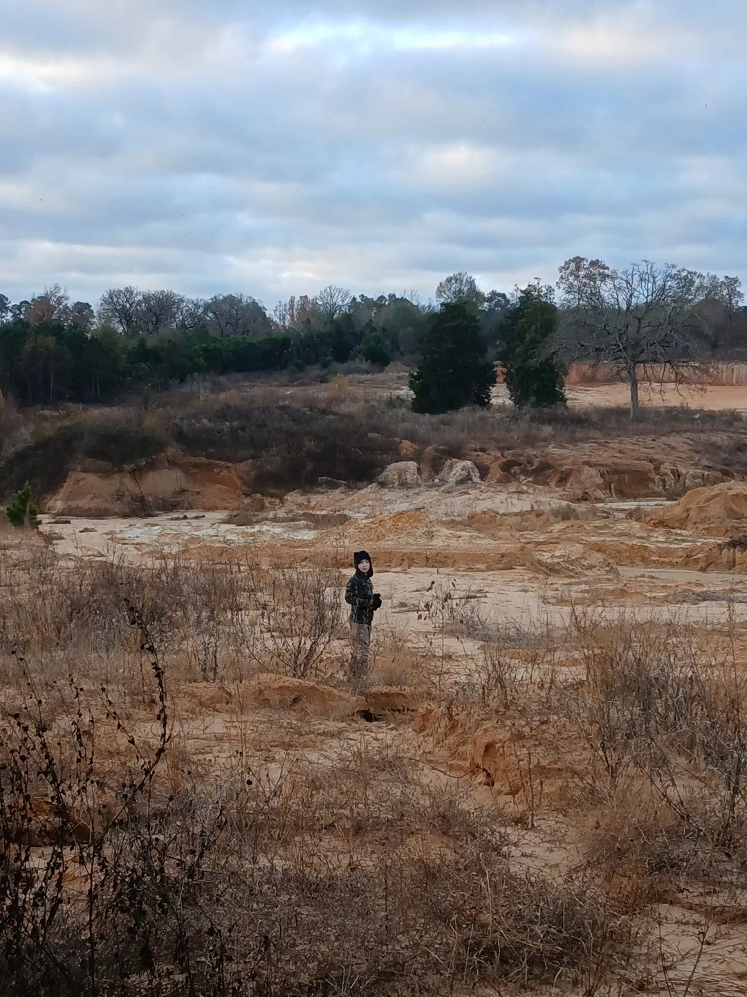 A person standing in a dry, barren landscape with sparse vegetation and a cloudy sky overhead.