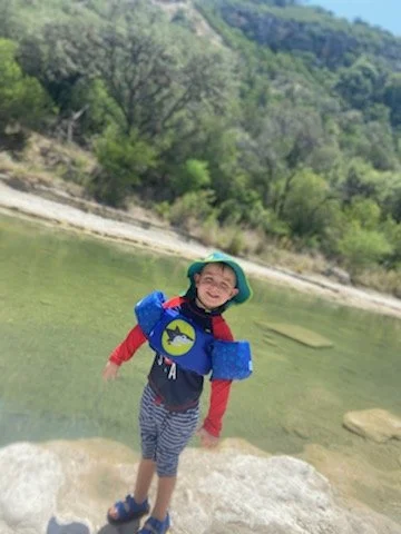 Child standing on a rock near a lake with trees and hills in the background, wearing a life jacket and hat.