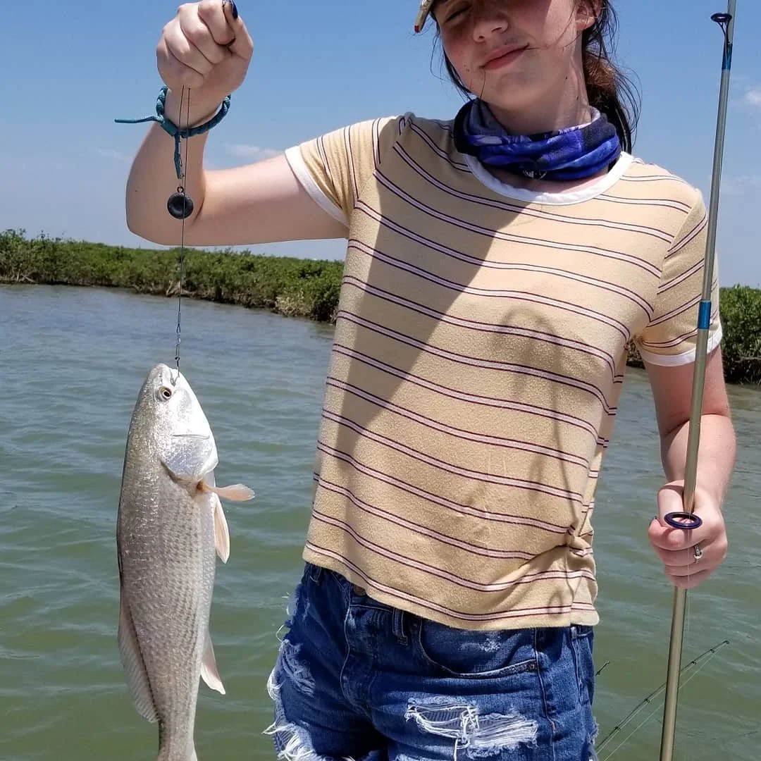 A young woman holding a fishing line with a caught fish in a body of water, standing outdoors with vegetation in the background.