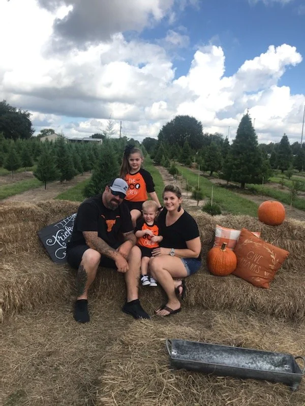 A family of four sitting on hay bales in a pumpkin patch with small pumpkins and fall-themed pillows nearby. The background features rows of evergreen trees under a partly cloudy sky.