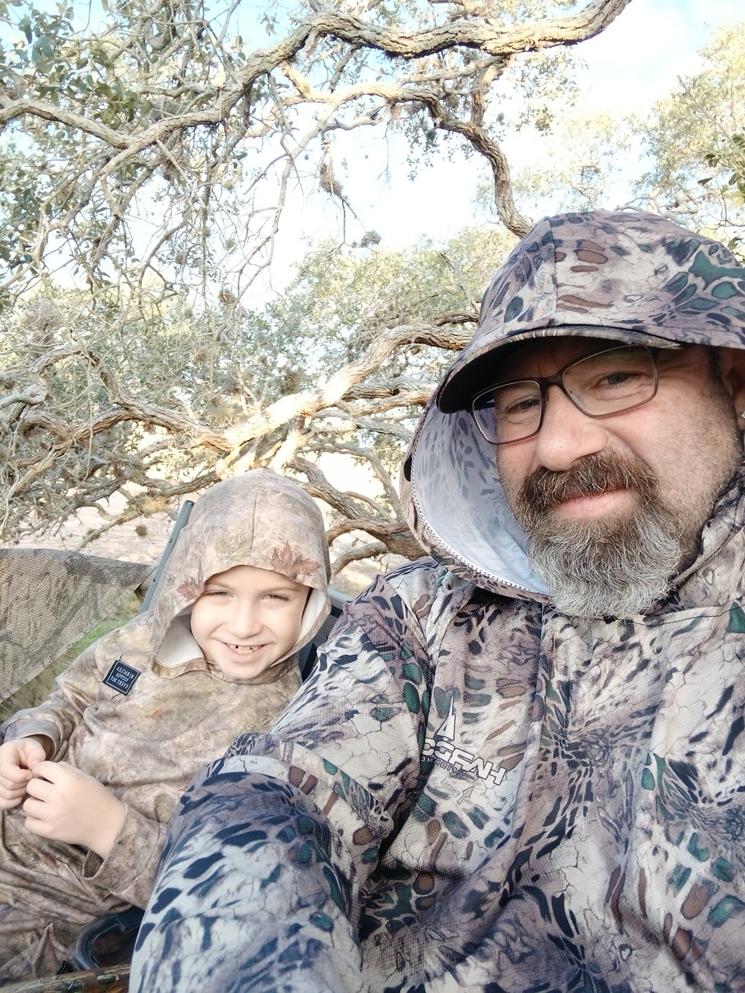 A man and a young boy outdoors in camouflage clothing, seated on a tree branch, with a background of twisted tree branches and light-colored sky.