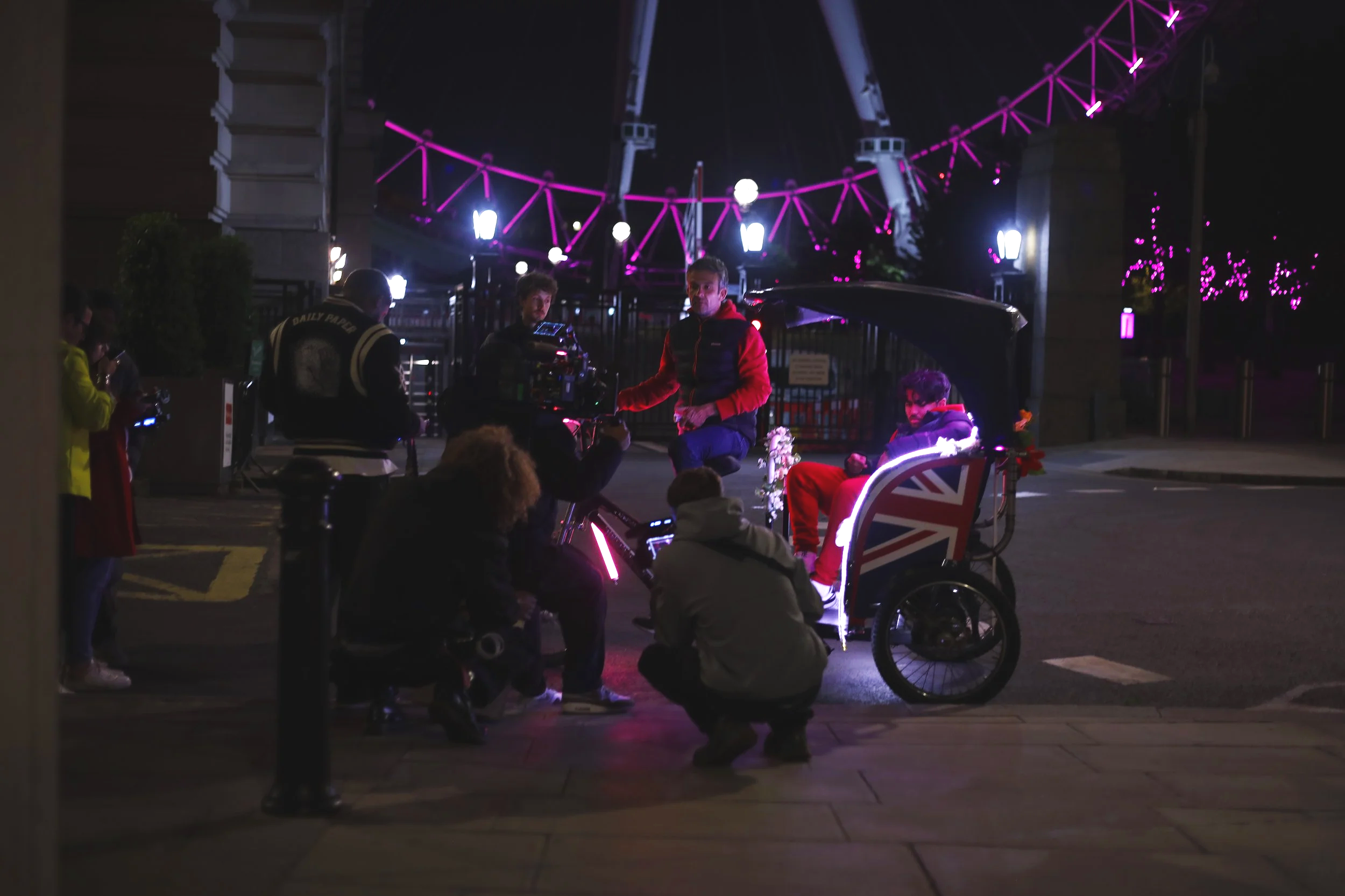 Nighttime scene featuring people gathered near a lit-up rickshaw with a Union Jack design, in front of a roller coaster illuminated with pink and purple lights.