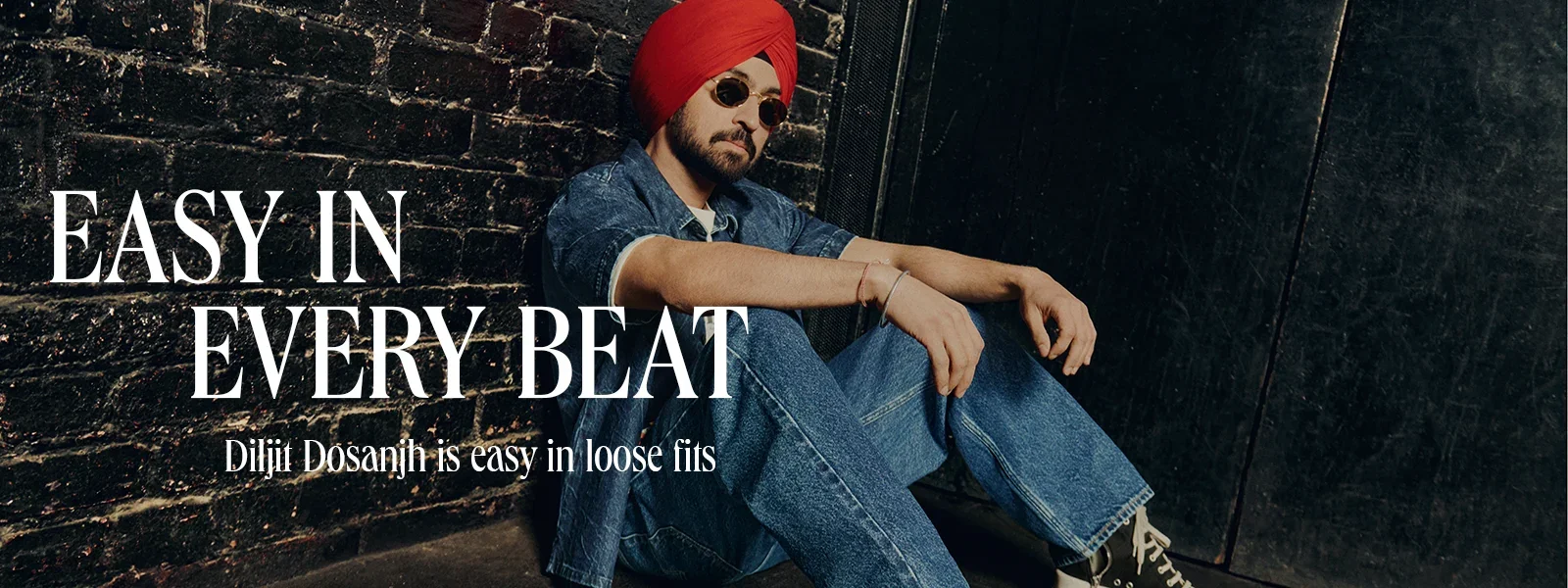 A man wearing sunglasses and a red turban, sitting on the floor against a black brick wall, dressed in a denim jacket and jeans, with white sneakers. Overlaid text reads 'EASY IN EVERY BEAT' and 'Diljit Dosanjh is easy in loose fits.'