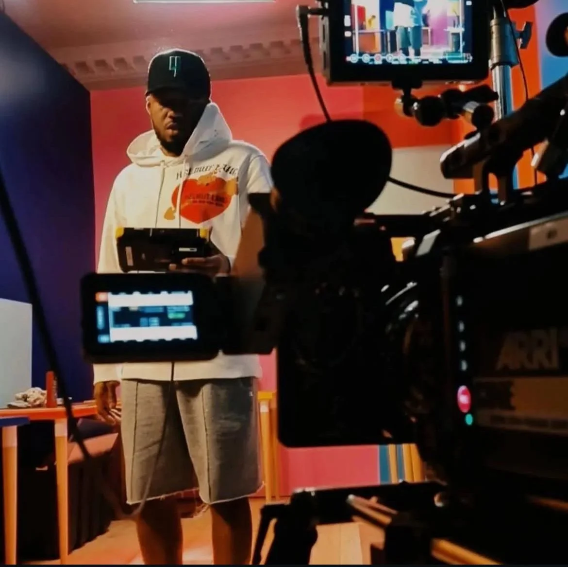 A man wearing a white hoodie and gray shorts holding a remote control, standing in front of a camera on a film set with colorful walls and a monitor showing a scene.