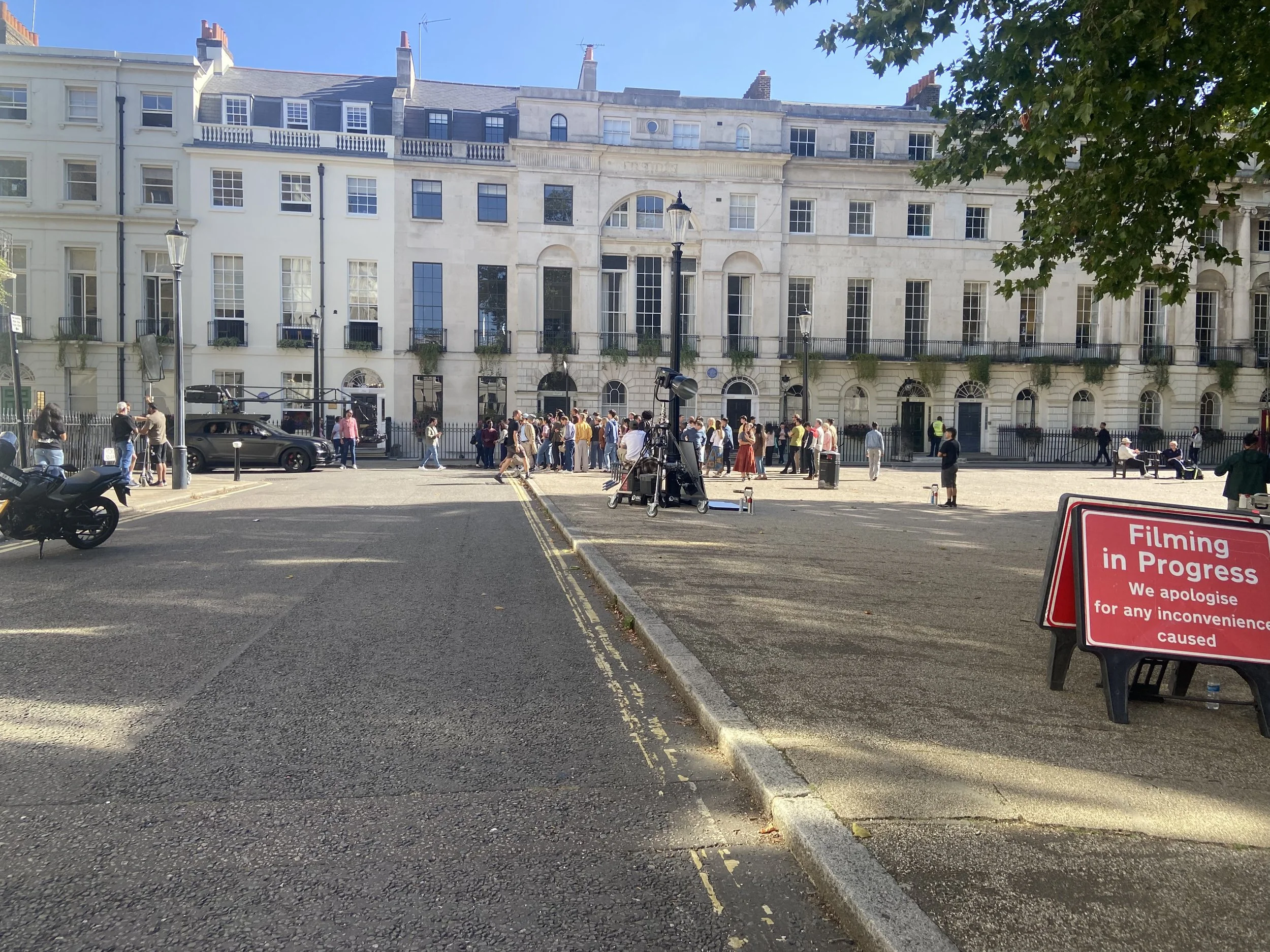 A street scene with a large, historic white stone building in the background, crowded with people walking and standing in front, some filming, as indicated by a red sign that reads 'Filming in Progress'. There are street lamps, parked cars, and motorcycles, with a clear blue sky overhead.