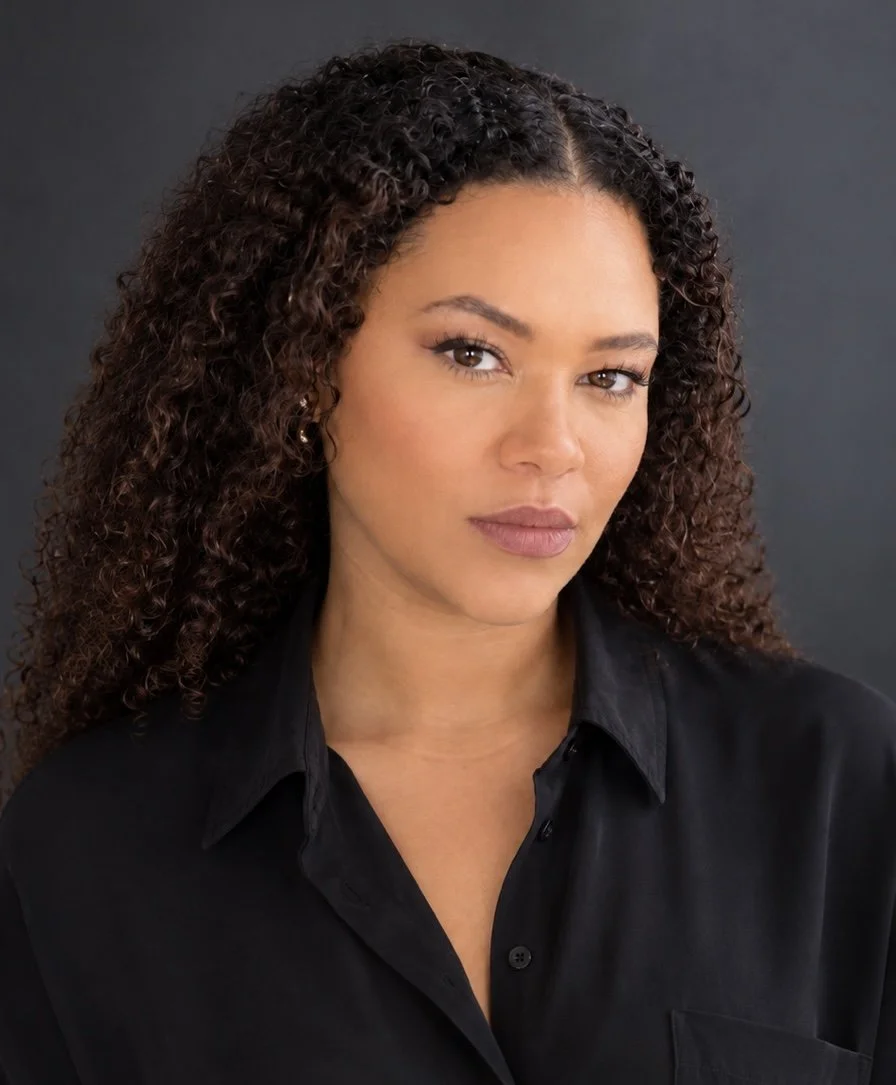 Close-up portrait of a woman with curly brown hair, light makeup, wearing a black button-up shirt, against a dark background.