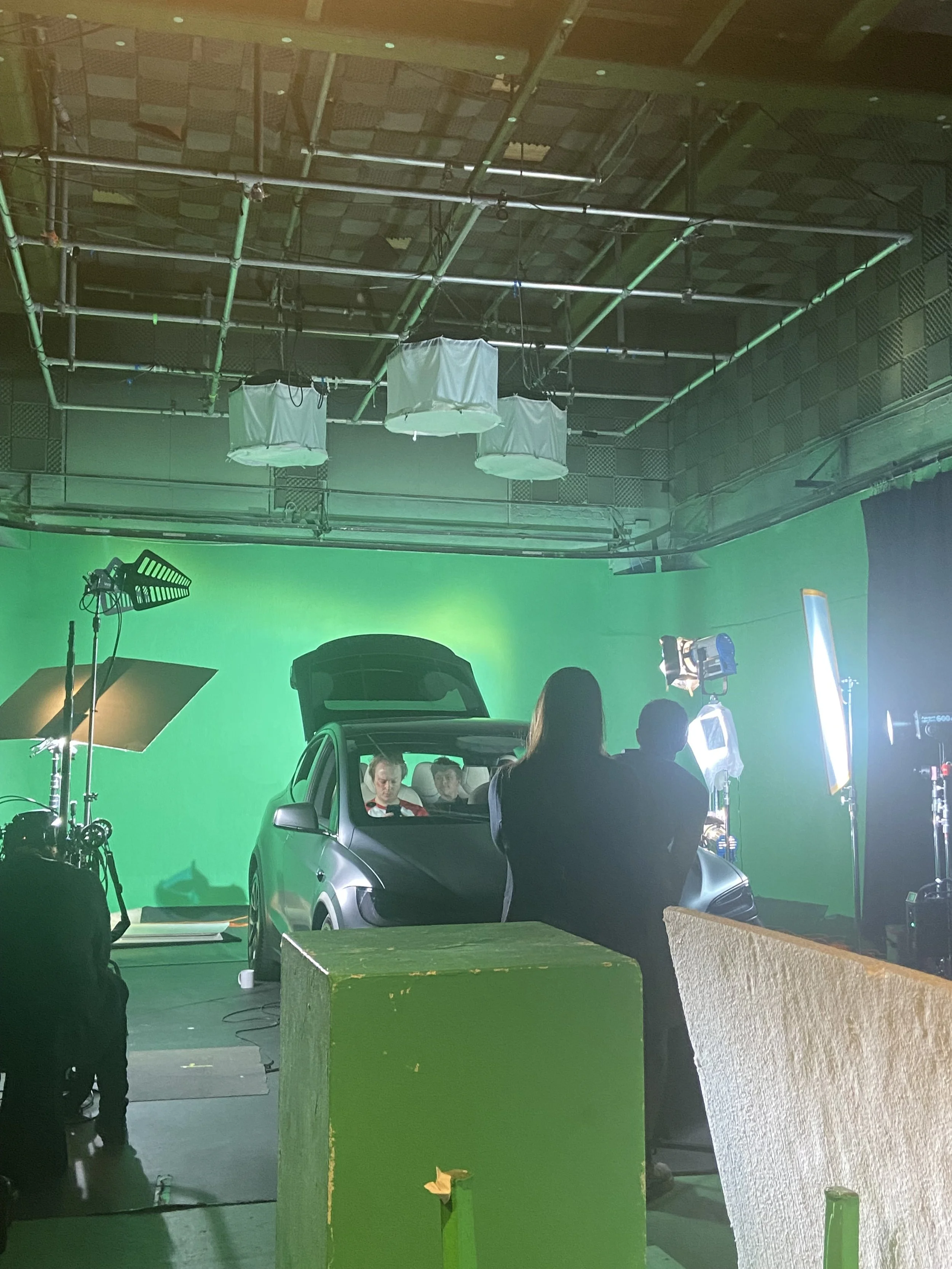 Behind-the-scenes view of a film or photo shoot set with a green screen, lighting equipment, a car, and crew members preparing for filming.