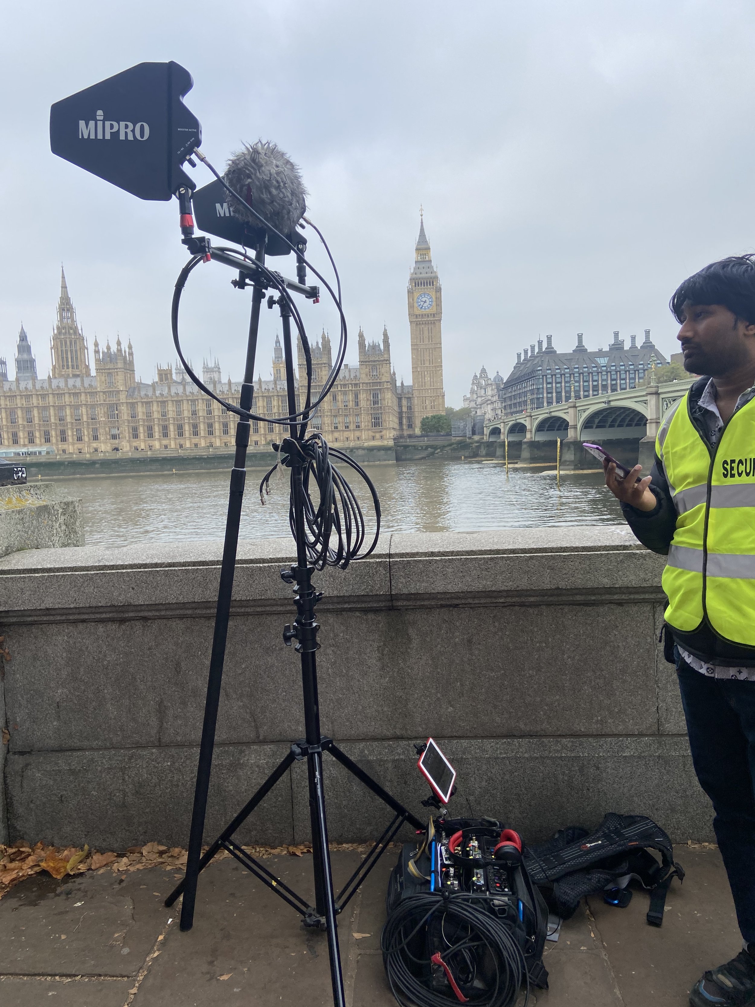 Audio and video recording equipment set up by a river, with the Big Ben clock tower and the Houses of Parliament in the background, and a man wearing a security vest holding a smartphone.