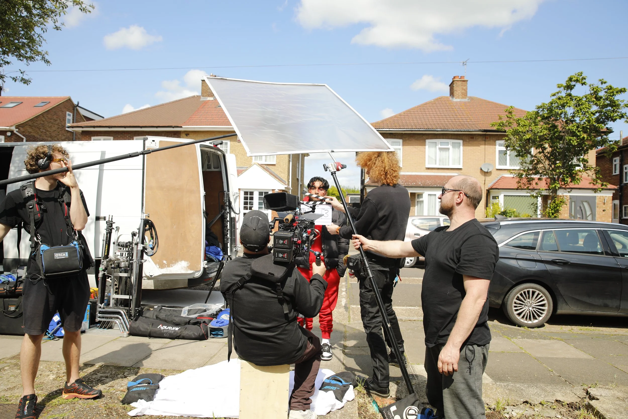 Filmmakers on a movie set outdoors, with equipment including a camera, boom microphone, and reflectors, in a residential neighborhood with brick houses and parked cars.