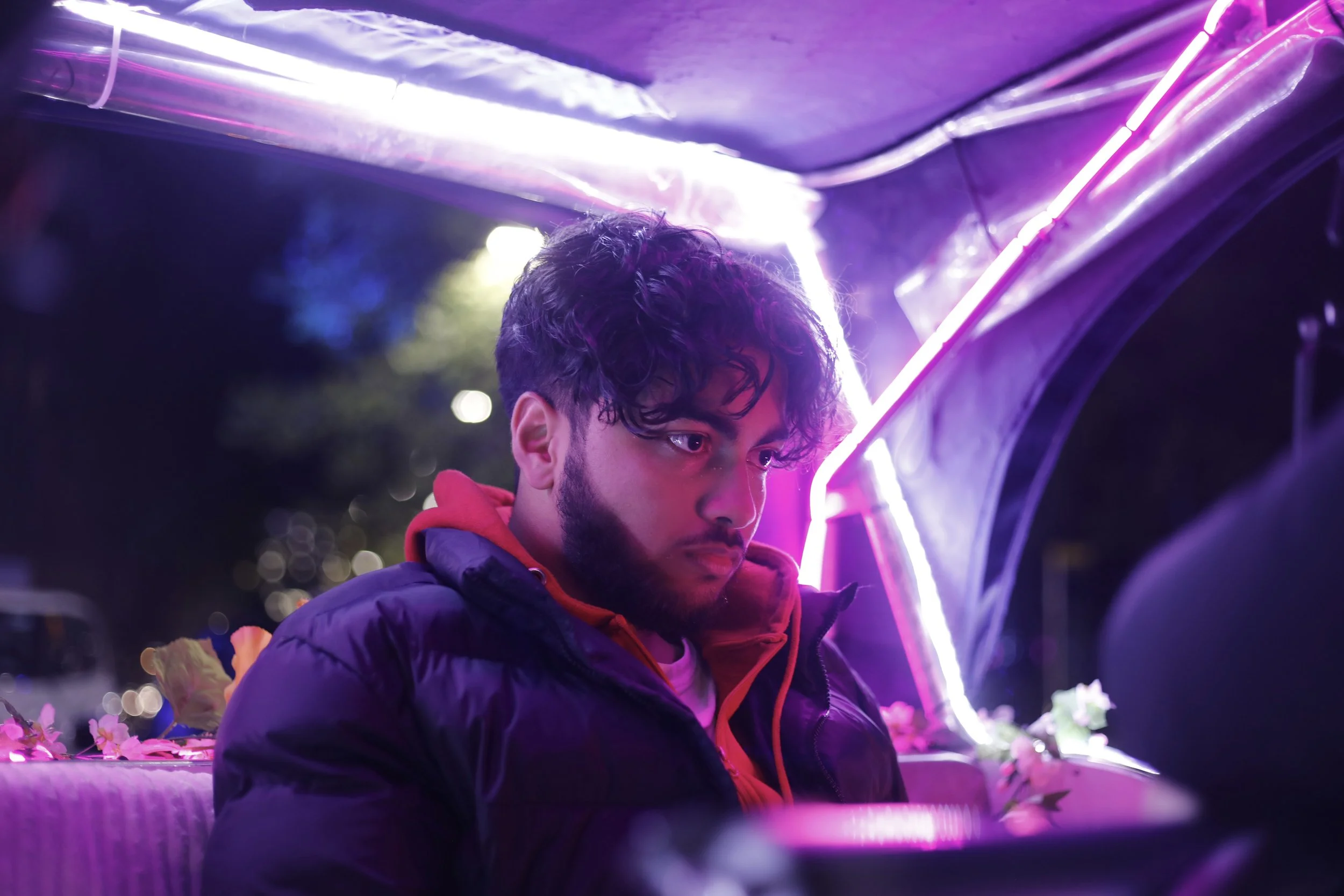 A young man with dark curly hair and a beard, wearing a red hoodie and a black jacket, sitting under vibrant purple and pink neon lights in a nighttime setting.