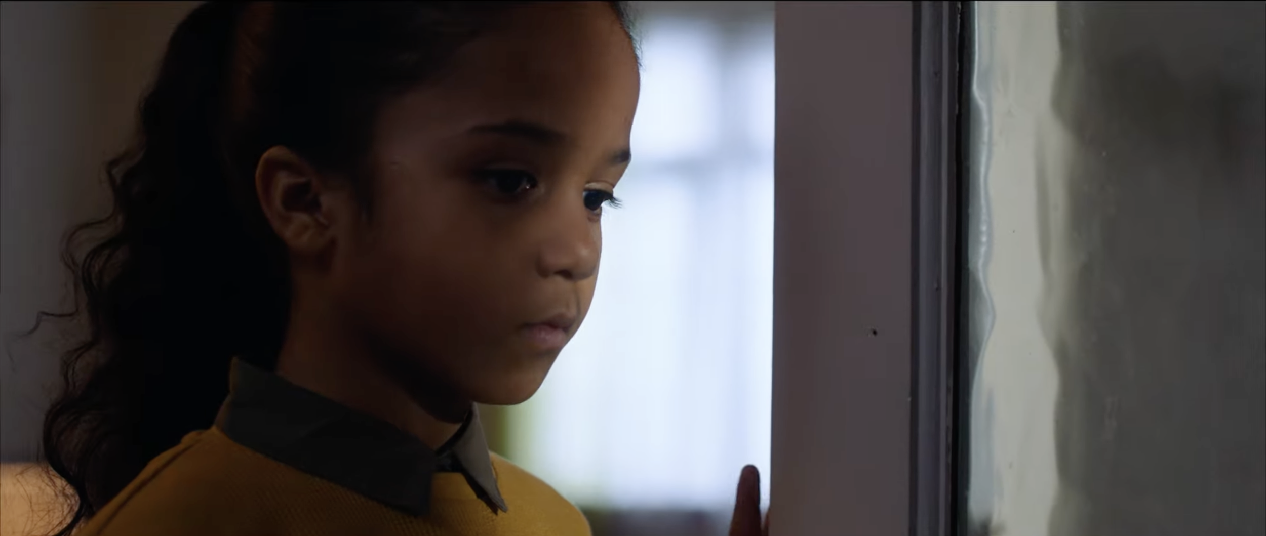 A young girl with curly hair looking at a window with melted snow or ice.