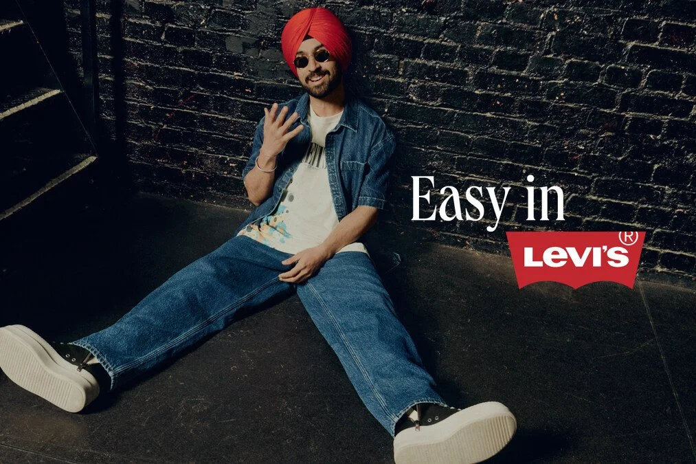 A man with a red turban and sunglasses sitting on the floor against a dark brick wall, wearing a denim jacket, ripped jeans, and sneakers, smiling and making a peace sign. The text reads "Easy in LEVI'S" with the Levi's logo.