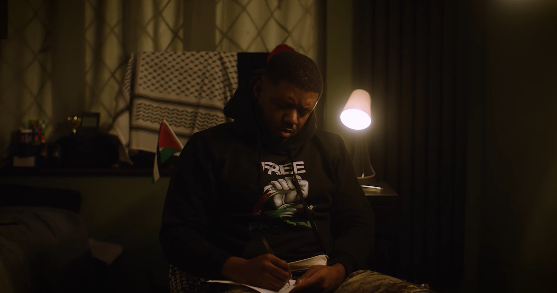 A young man sitting on a bed in a dimly lit room, writing in a notebook, wearing a hoodie and a t-shirt with the words "Free" on it; there is a desk with a small flag, a bell, and a Rubik's cube in the background, with a lamp illuminating the scene.