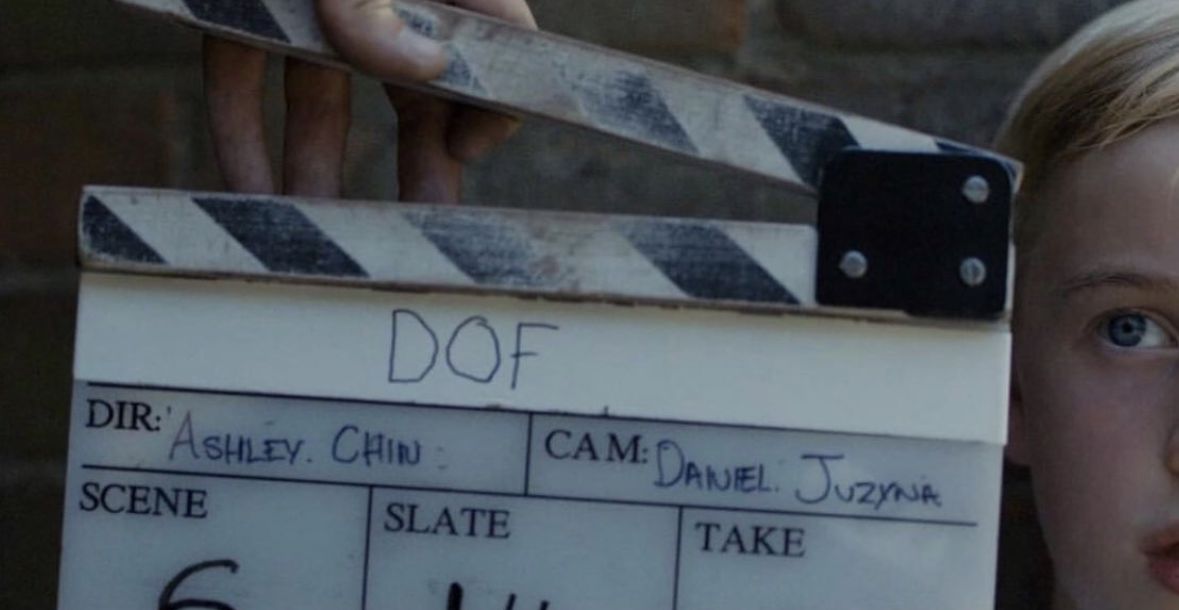A film slate with handwritten notes, held by a person, partially showing a child's face with blonde hair and blue eyes.