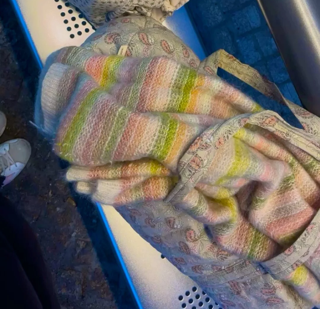 Ein bunt gestrickter Mohair Pullover in Grün- und Rosatönen, liegend auf einer Reisetasche auf einer Bank 